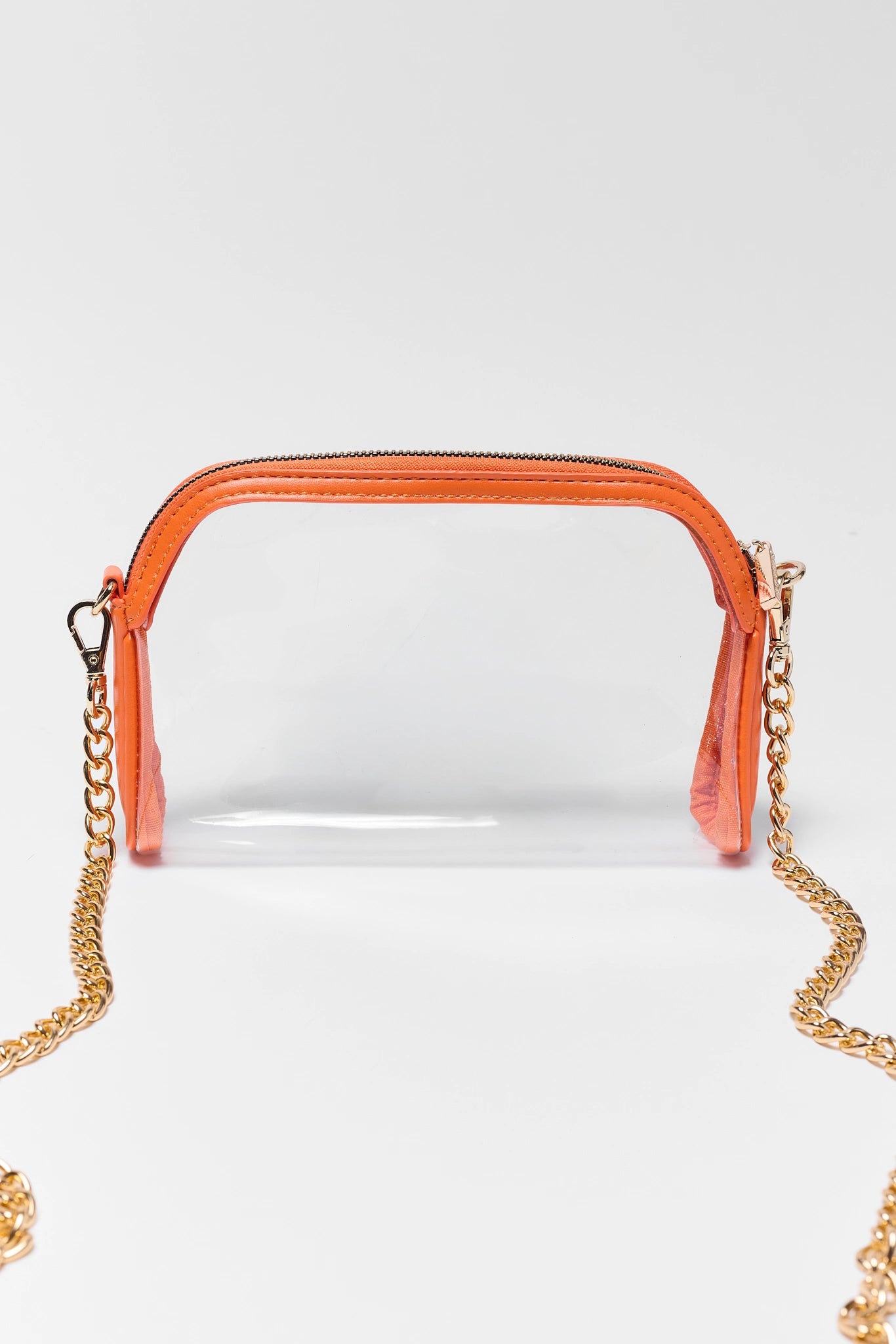 A clear acrylic crossbody bag with a bright orange top handle and trim, featuring a gold-tone chain strap, photographed against a light gray studio background.