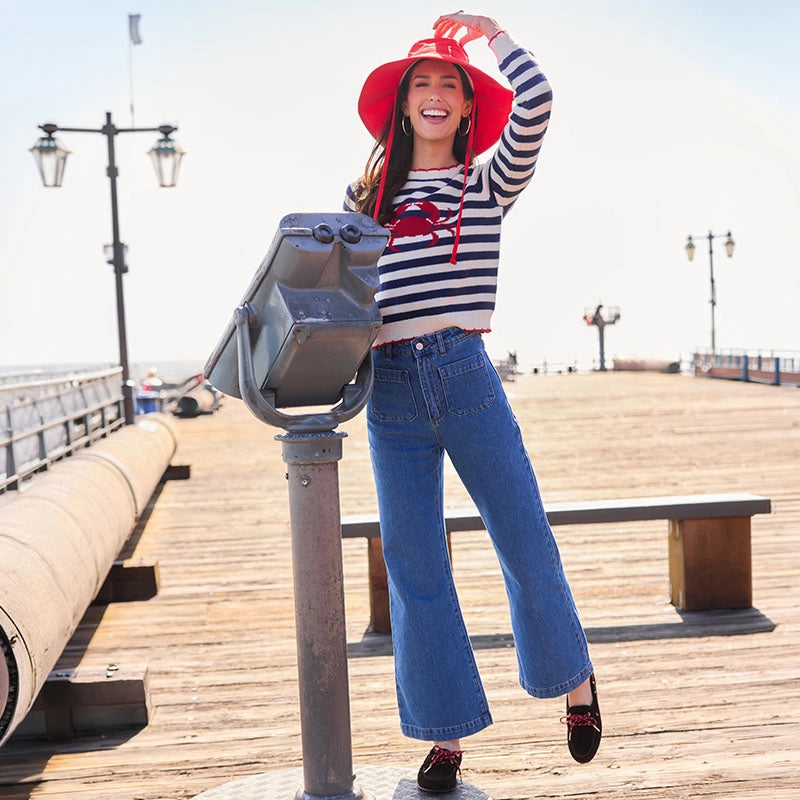 Woman in a striped shirt and red hat standing on a wooden pier with a bench and lamp post in the background.