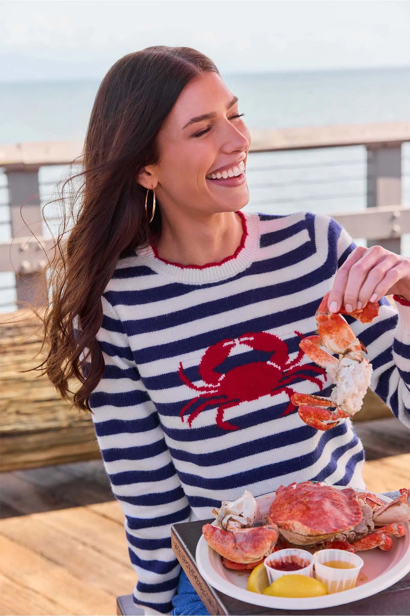 Woman in a striped sweater with a crab design, holding seafood by the water.