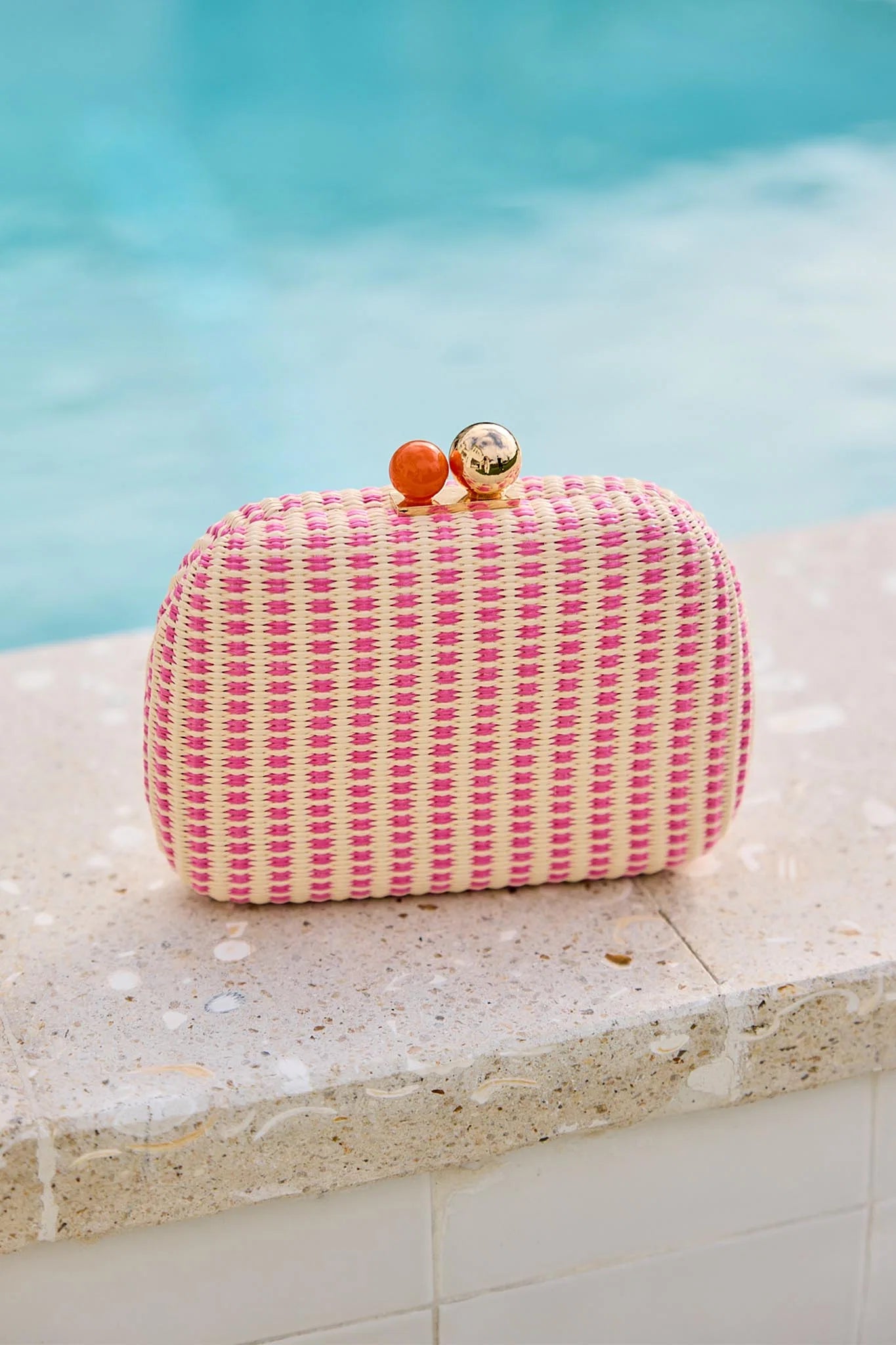 Pink woven clutch with cherry decorations on a stone ledge by a pool.
