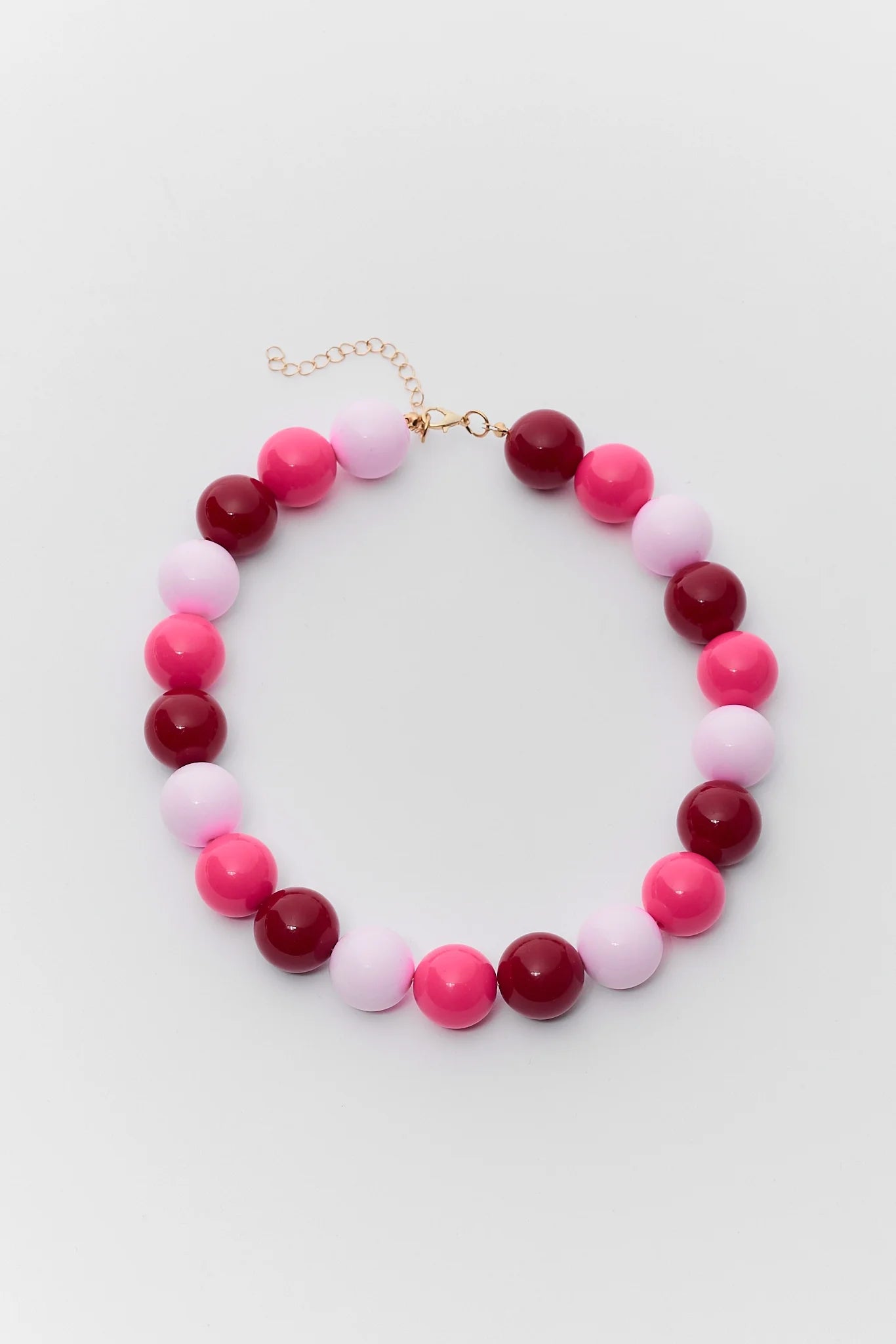 A beaded necklace with alternating glossy pink, deep magenta, and pale pink round beads, finished with a gold-tone clasp and extender chain, shown on a light gray background.