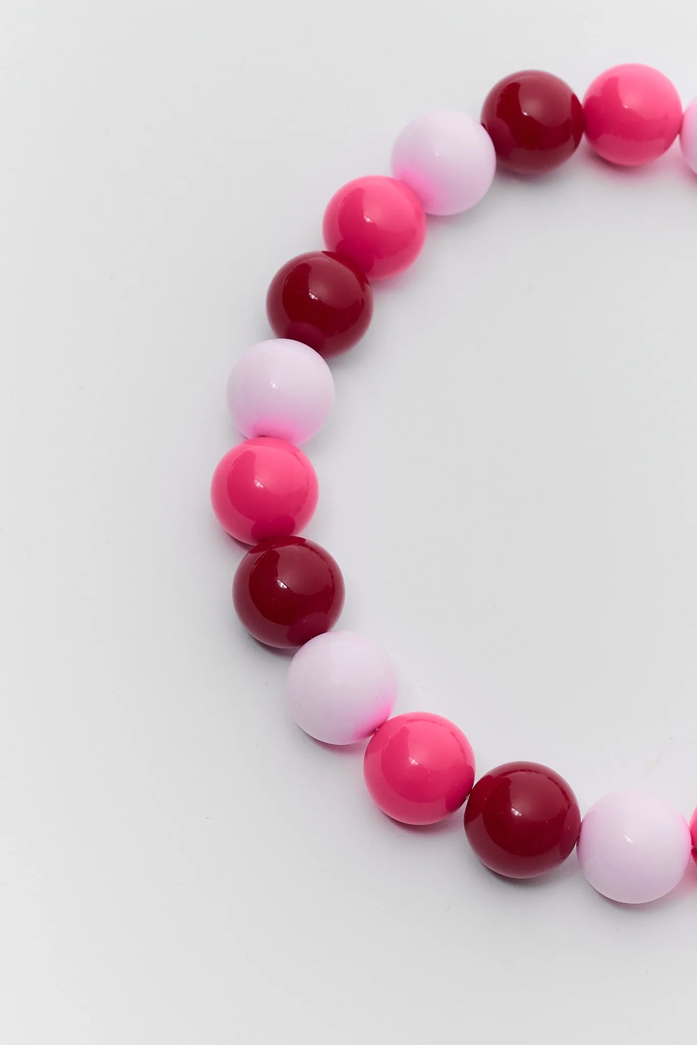 A beaded stretch bracelet made of glossy round beads in alternating shades of pink, magenta, and deep red, arranged in a curve on a light gray background.