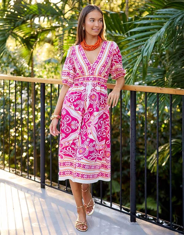 Woman in a pink and white patterned dress standing on a wooden deck with greenery in the background nav