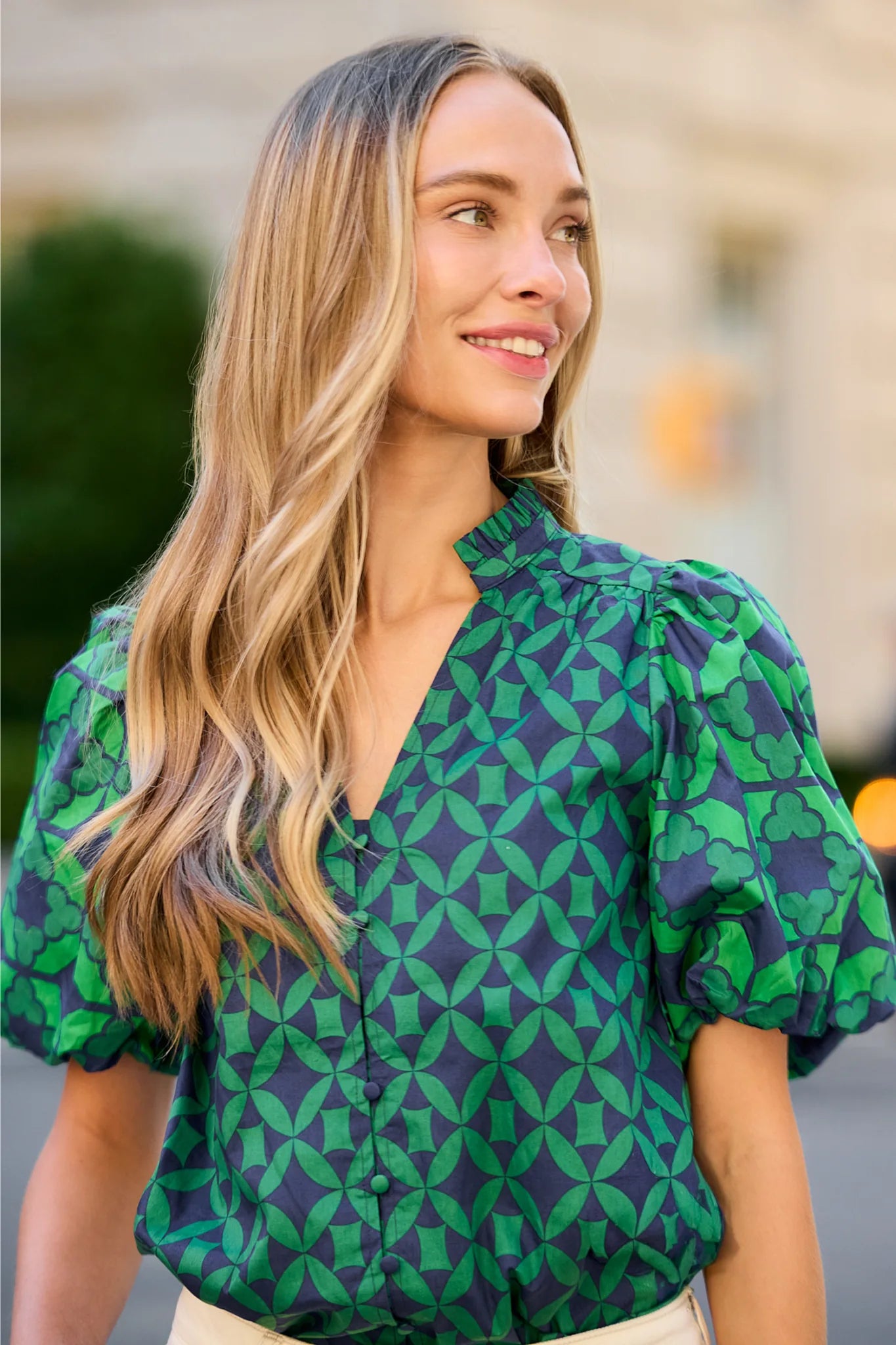 A woman with long blonde hair wearing a green and navy blue geometric patterned blouse with puff sleeves, photographed outdoors against a softly blurred background.