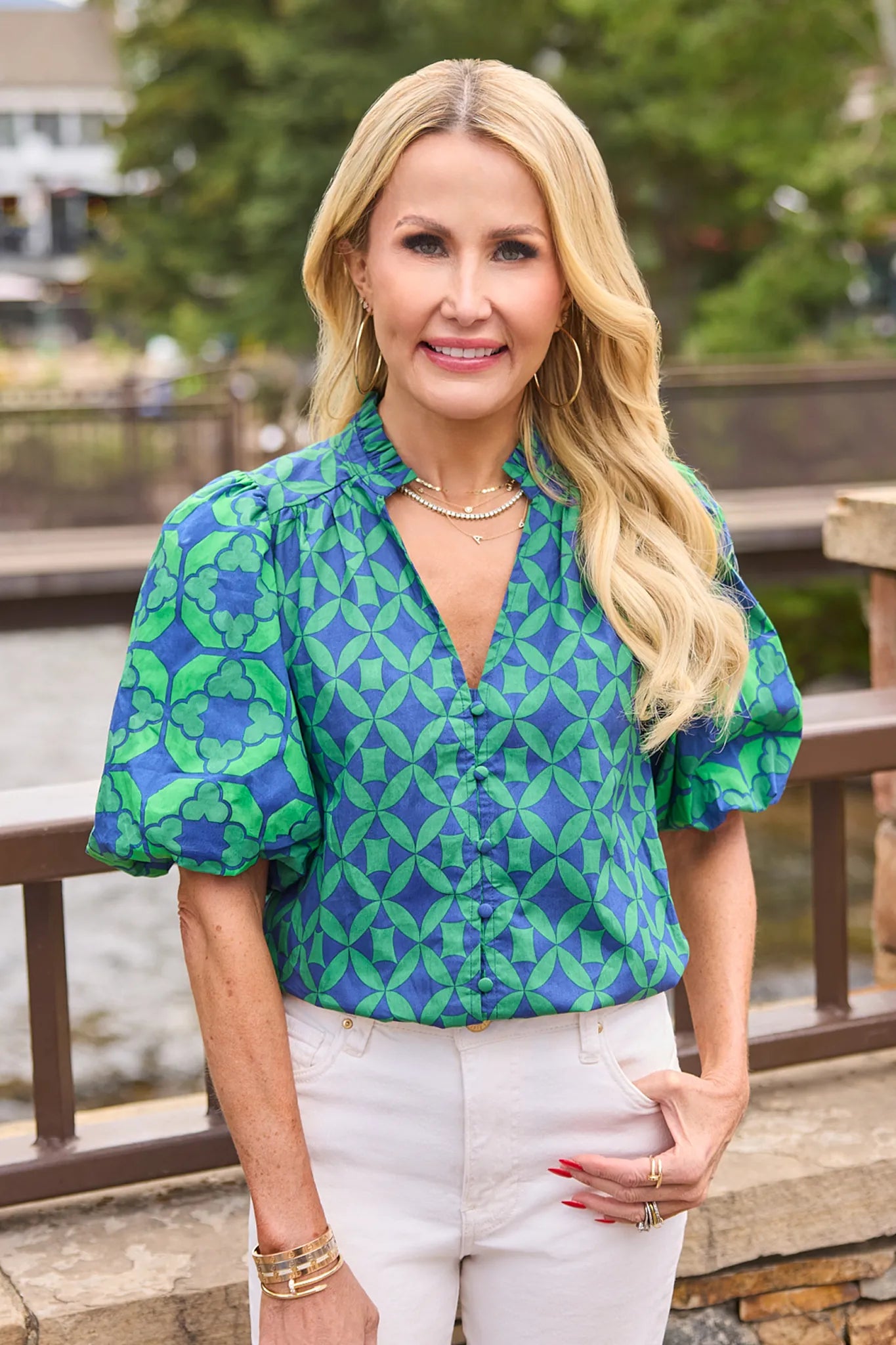 A blonde woman wearing a green and blue patterned short-sleeve blouse with puff sleeves and white pants, standing outdoors near a stone railing and water.