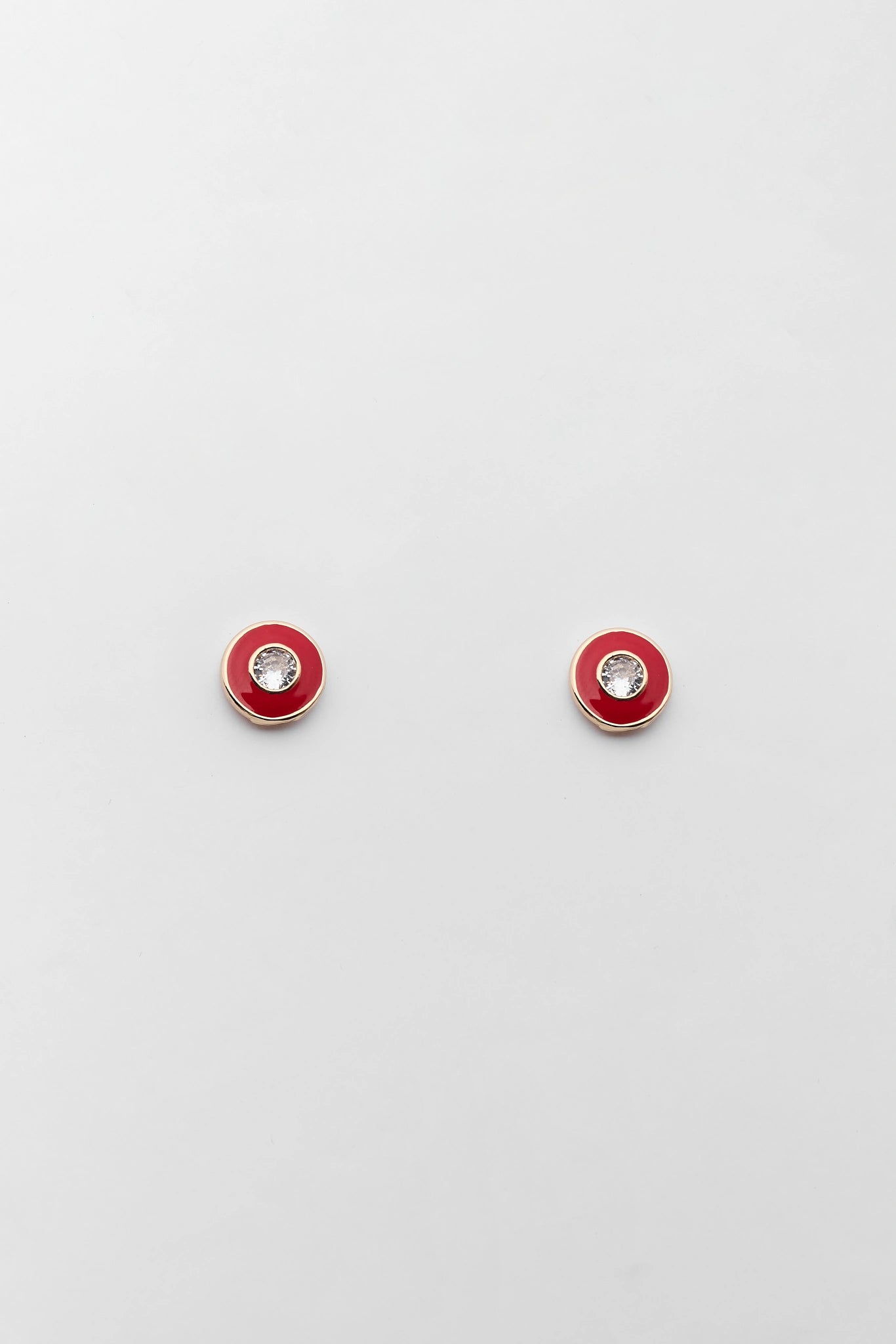 A pair of small round stud earrings with a vibrant red enamel face, a silver-toned metal rim, and a clear central gemstone, displayed on a clean white background.
