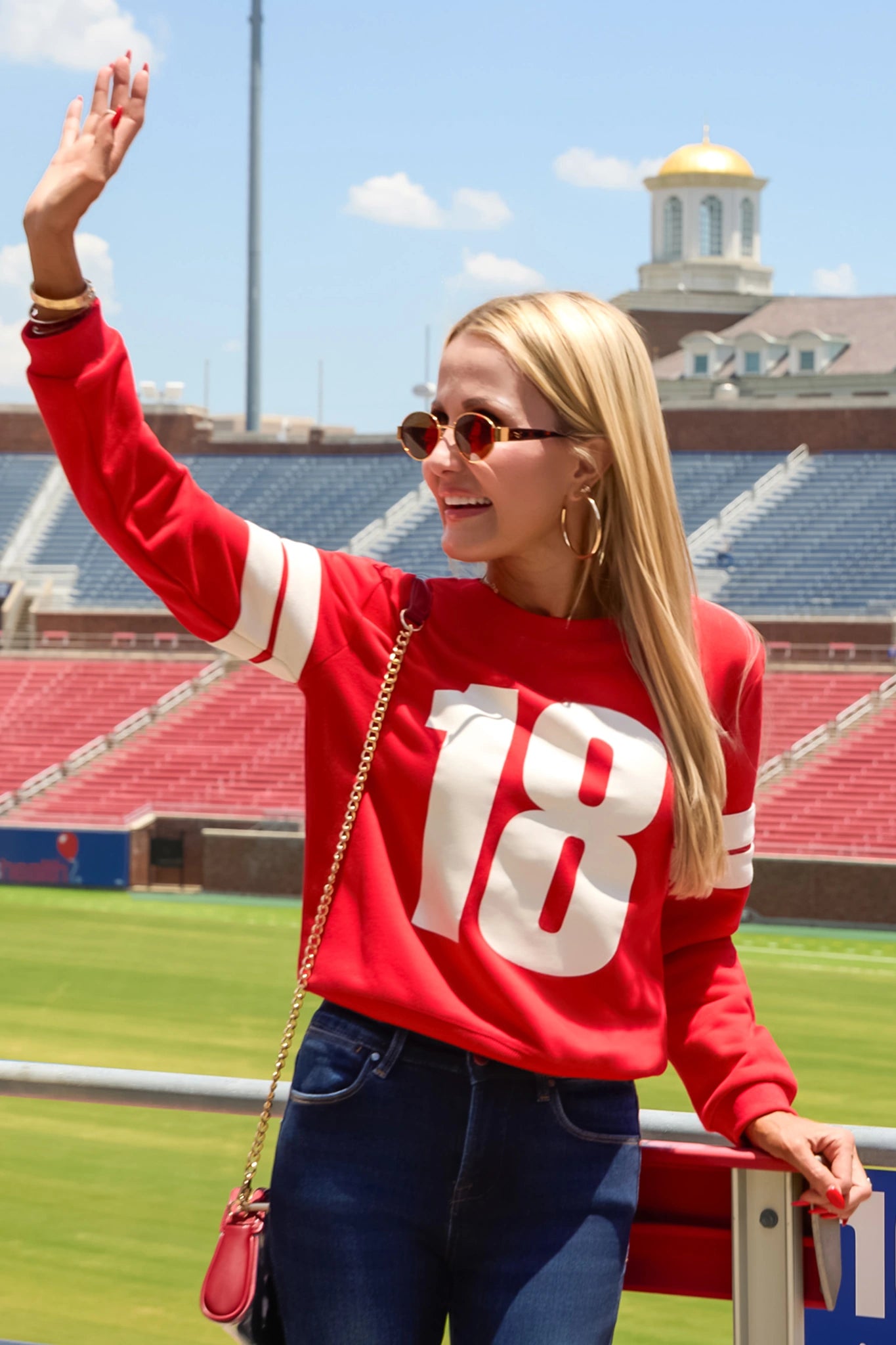 A woman standing on a sports field wearing a bright red crewneck sweatshirt with the number 18 in white on the front, paired with dark blue jeans and a small red crossbody bag.