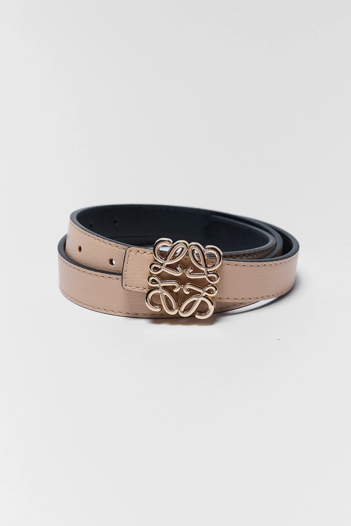 A light beige leather belt with a black interior lining and a gold-tone interlocking script logo buckle, coiled on a light gray background.