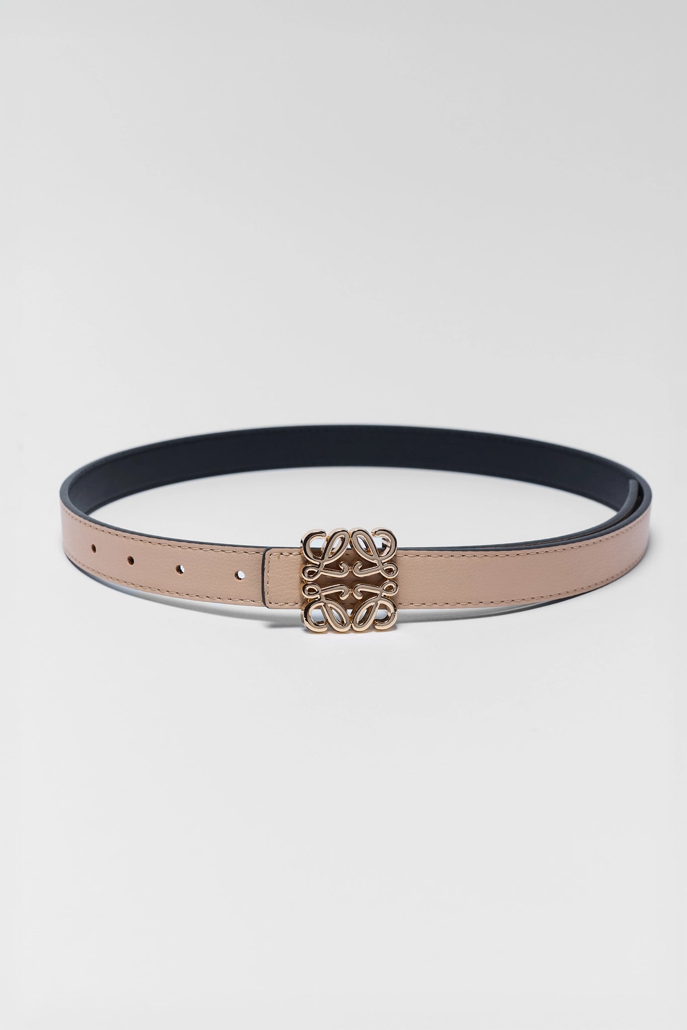 A slim tan leather belt with a gold-tone interlocking monogram buckle, displayed in a loop on a light gray background.