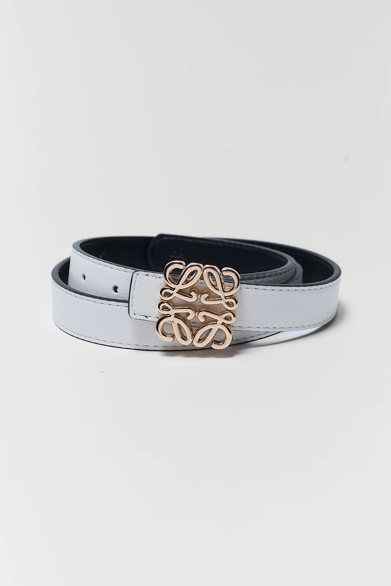 A light gray leather belt with a prominent interlocking monogram buckle in a warm gold-tone finish, coiled on a light gray background.