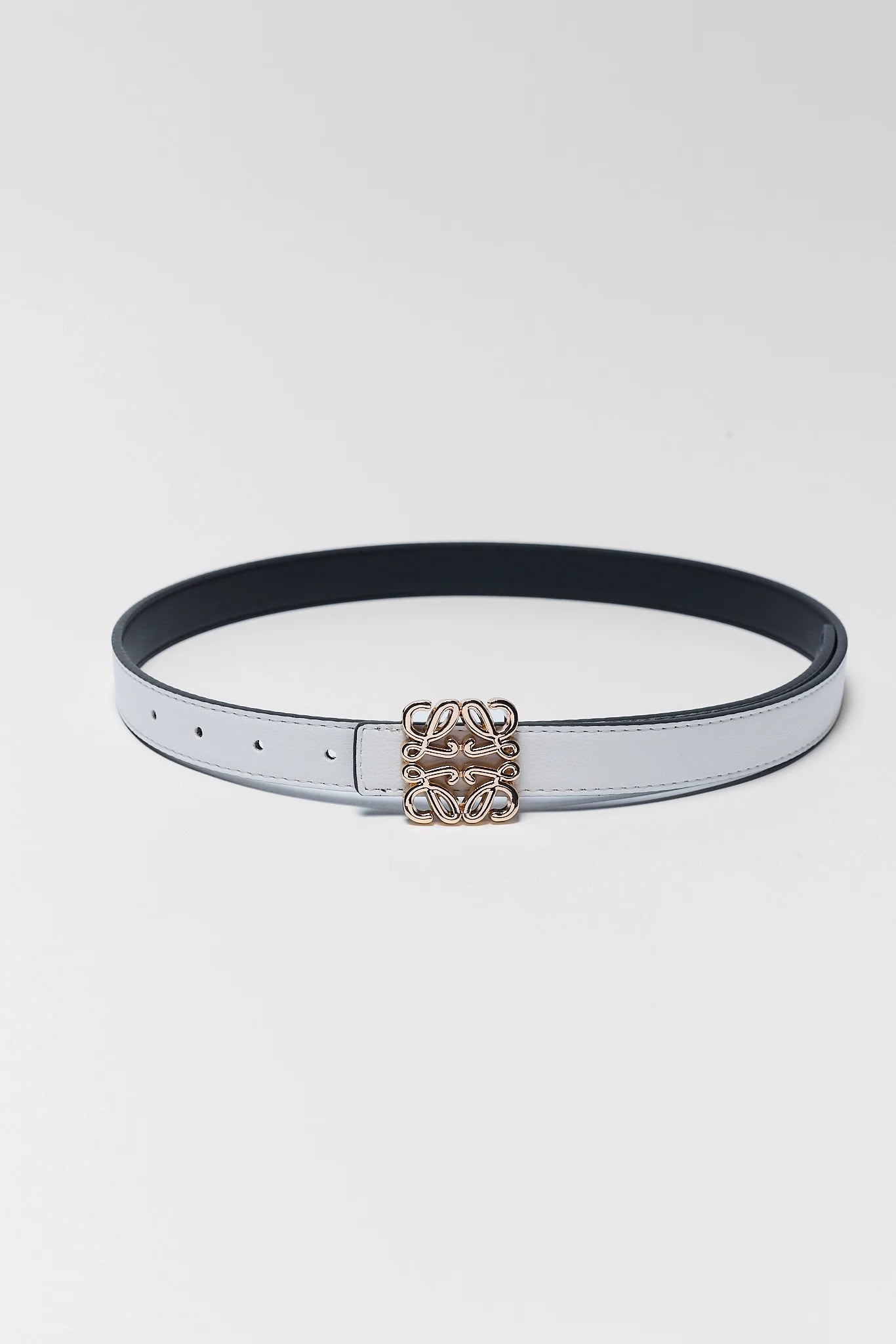 A slim white leather belt with a black underside and a gold-tone interlocking monogram buckle, displayed on a light gray background.