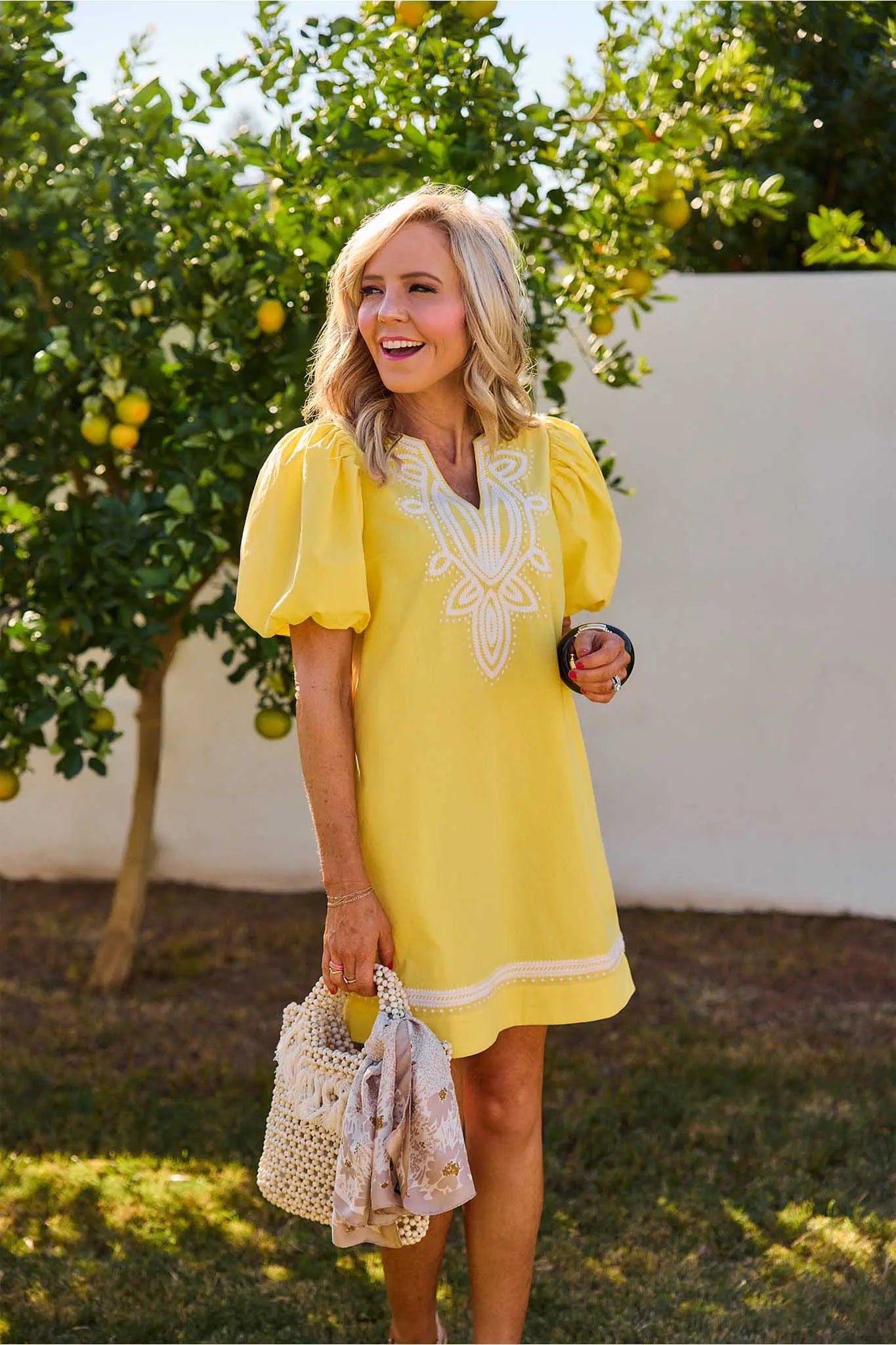Woman in a Roxanne Yellow Embroidered Dress with white embroidery standing outdoors near an orange tree.