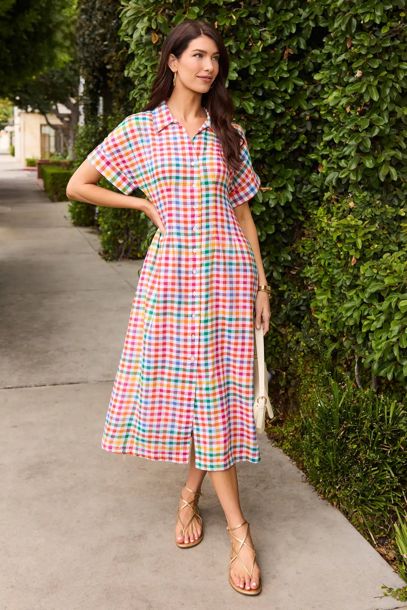 Woman wearing a colorful checkered dress standing on a sidewalk next to greenery.