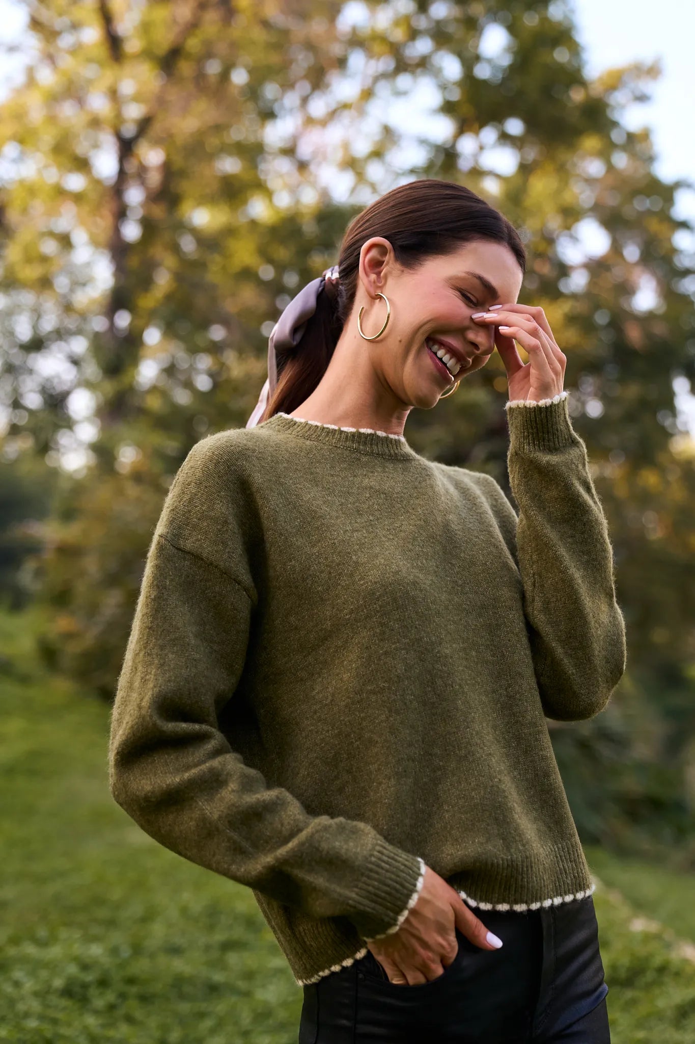 Woman wearing a olive Lottie Sweater with a blurred natural background