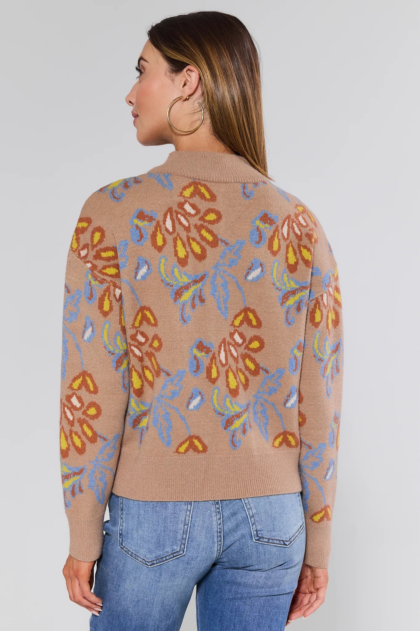 Woman wearing a beige Nancy Sweater with colorful floral patterns and blue jeans on a plain background