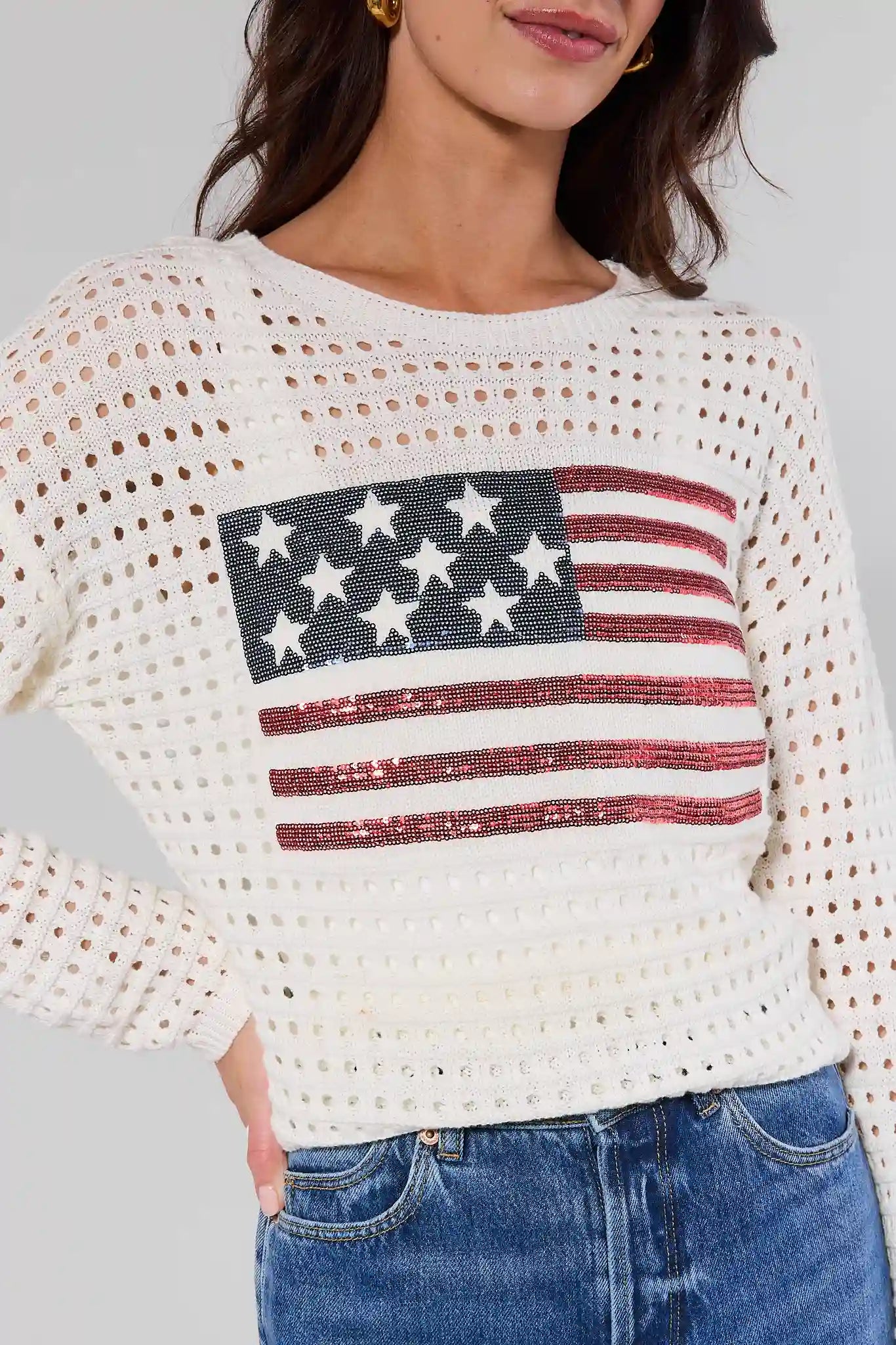 close up Person wearing a white crochet top with an American flag design and blue jeans.