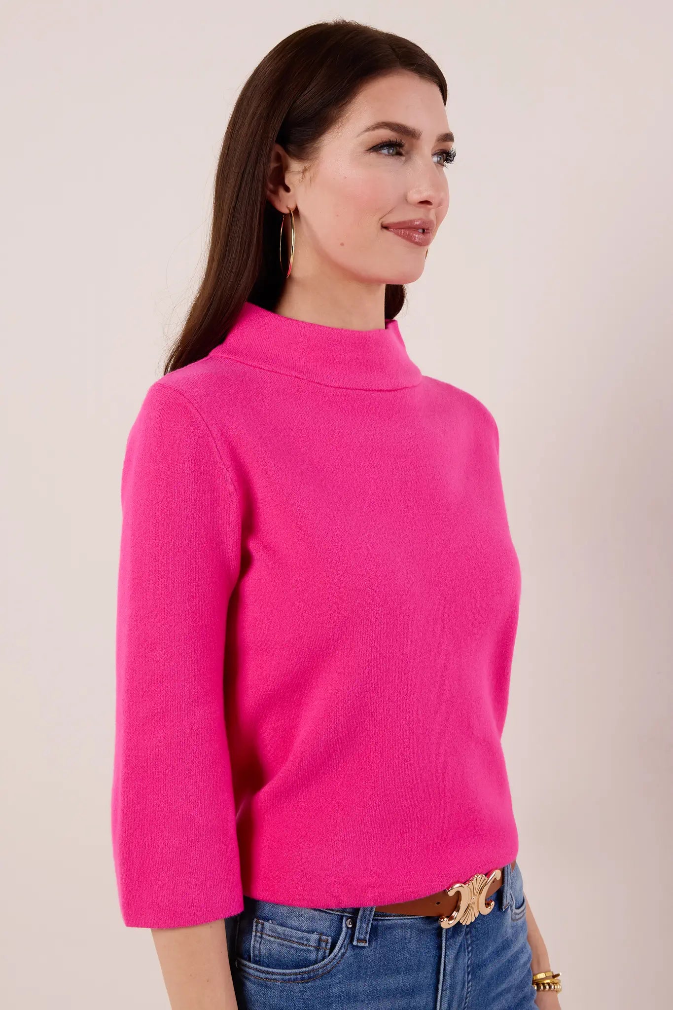 Woman wearing a bright pink Jackie sweater and blue jeans against a plain background
