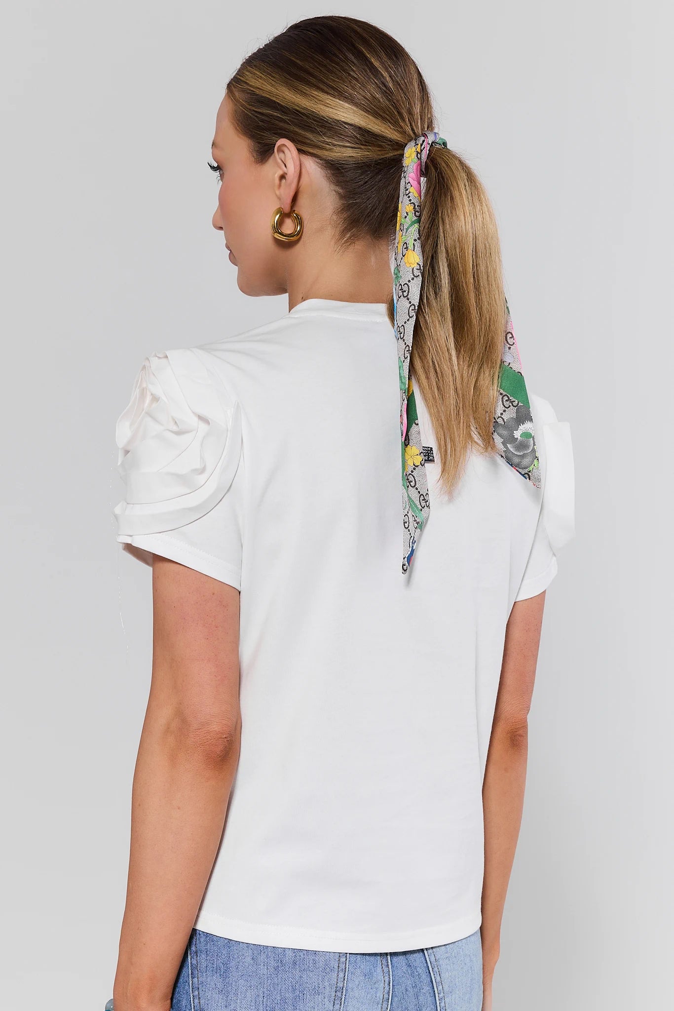 Woman standing wearing a white blouse with floral hair tie on a plain background