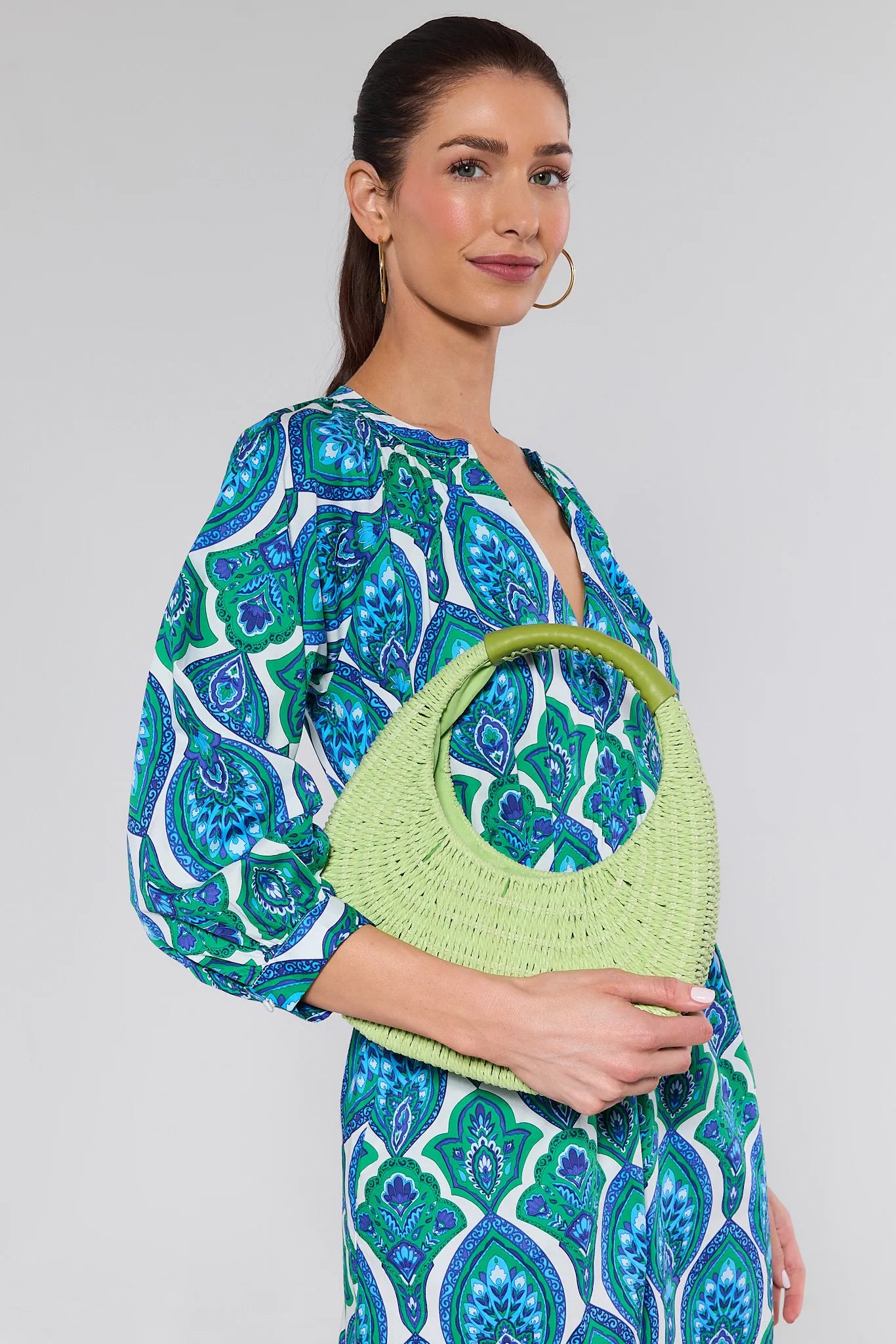 Woman holding a green handbag wearing a blue and green patterned dress on a gray background