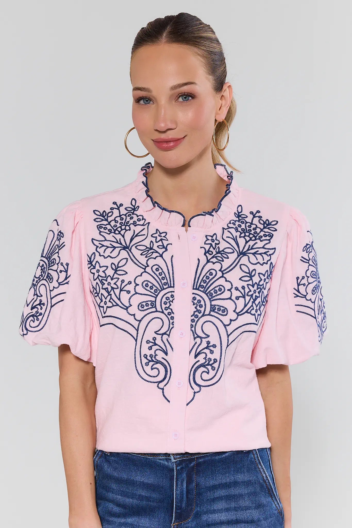 Woman wearing a pink blouse with blue embroidery on a plain background