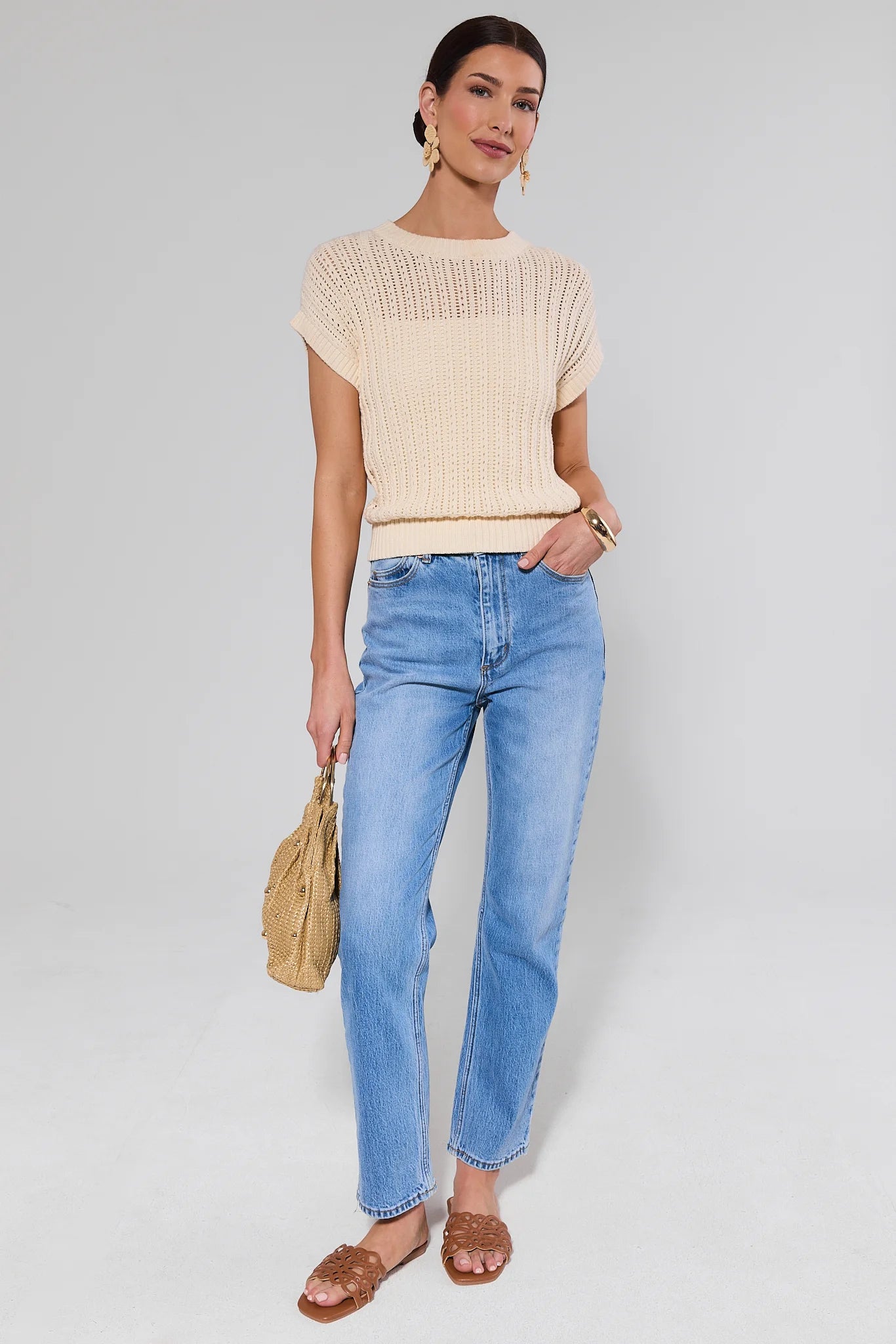 Woman wearing a beige top and blue jeans, holding a beige handbag on a light gray background