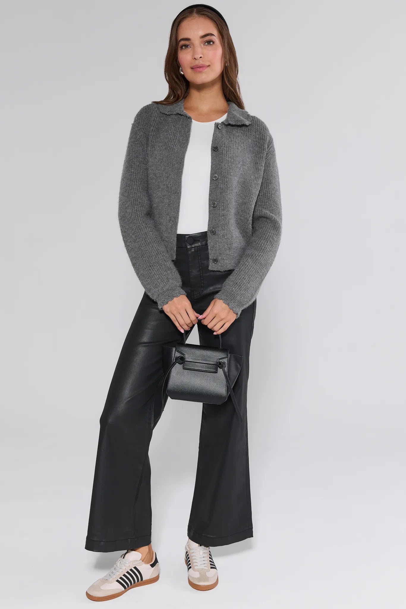 Woman wearing a gray cardigan, white shirt, black Meg Jeans Wax Coated, and sneakers on a white background