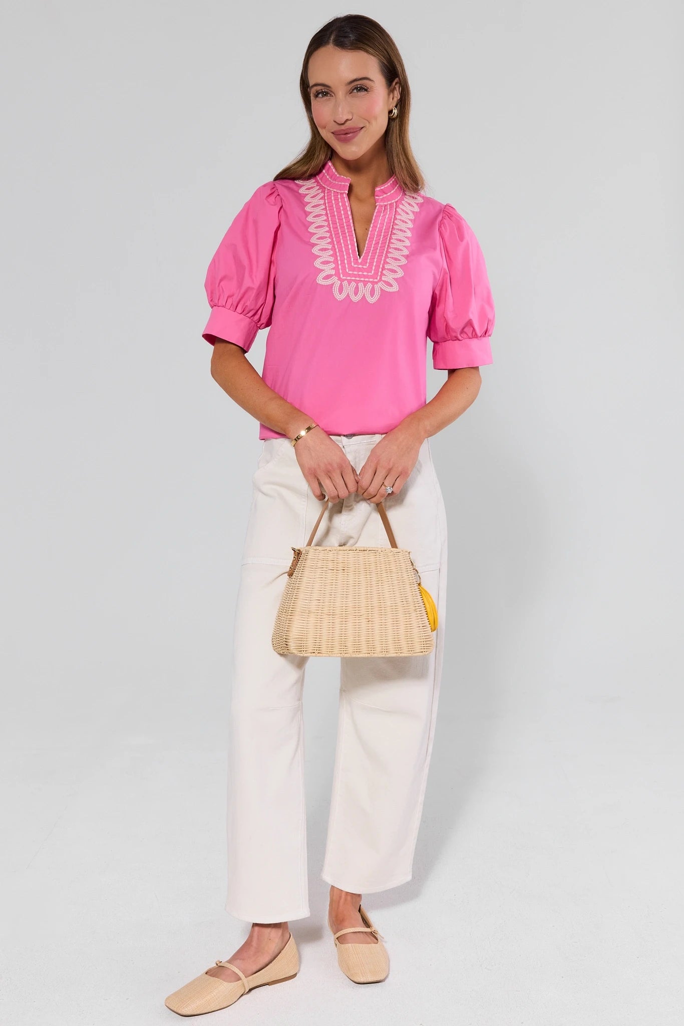 Woman wearing a pink blouse with white trim and white pants, holding a straw bag on a light gray background.