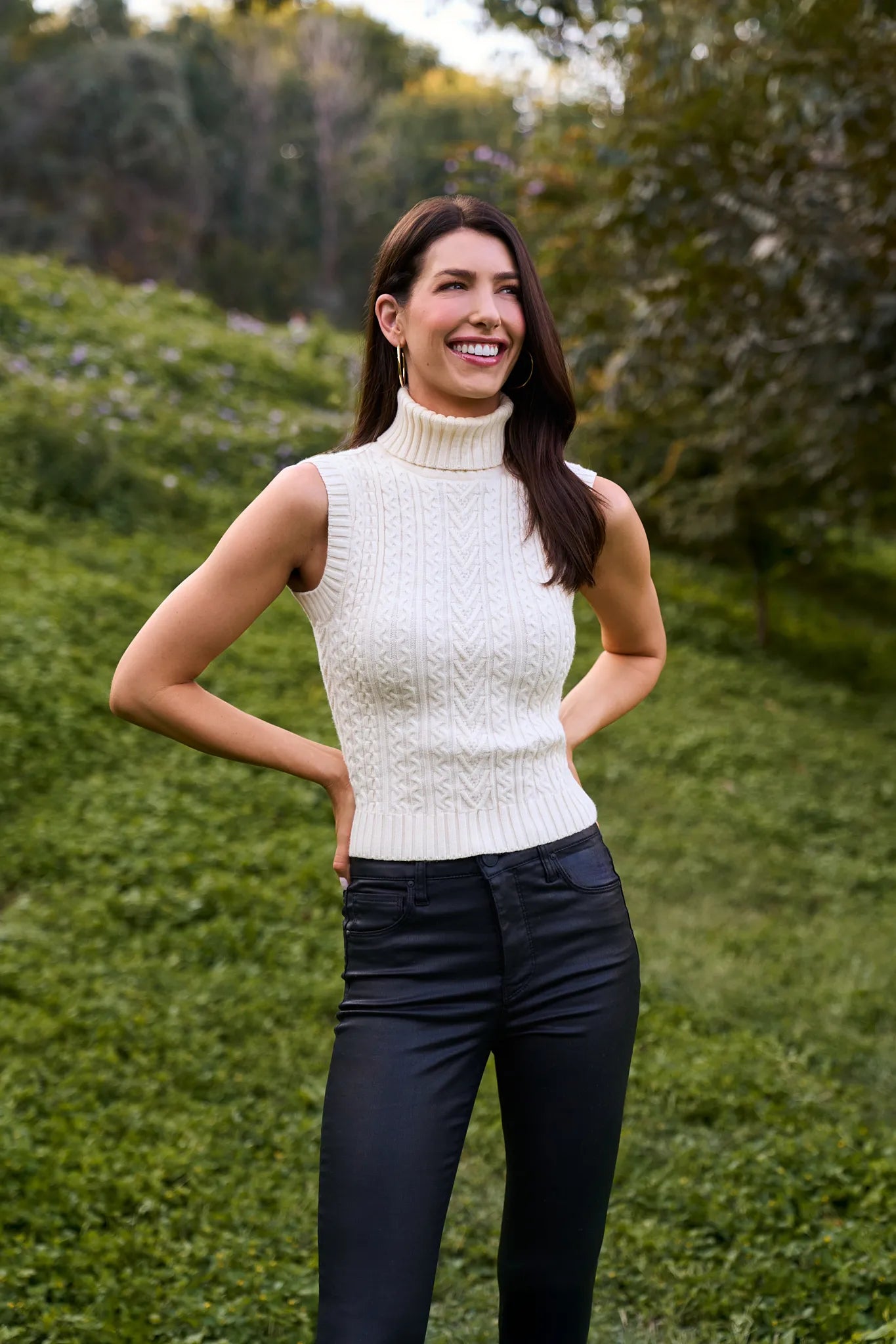 Woman wearing a cream sleeveless Sienna Sweater and dark pants standing outdoors.