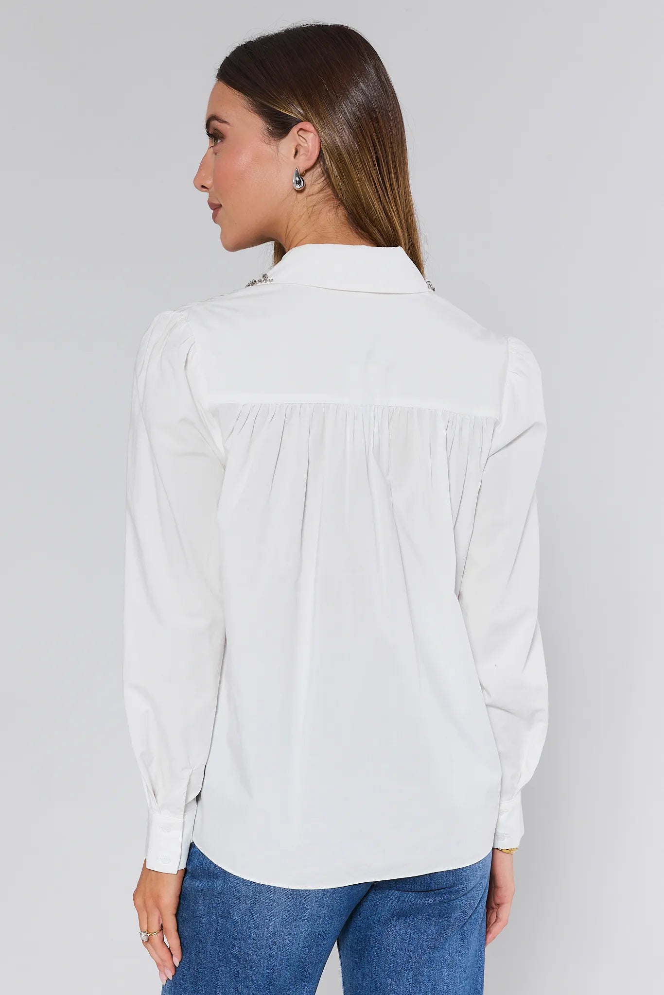 Woman wearing a white Celeste Top with embellishments and blue jeans on a plain background