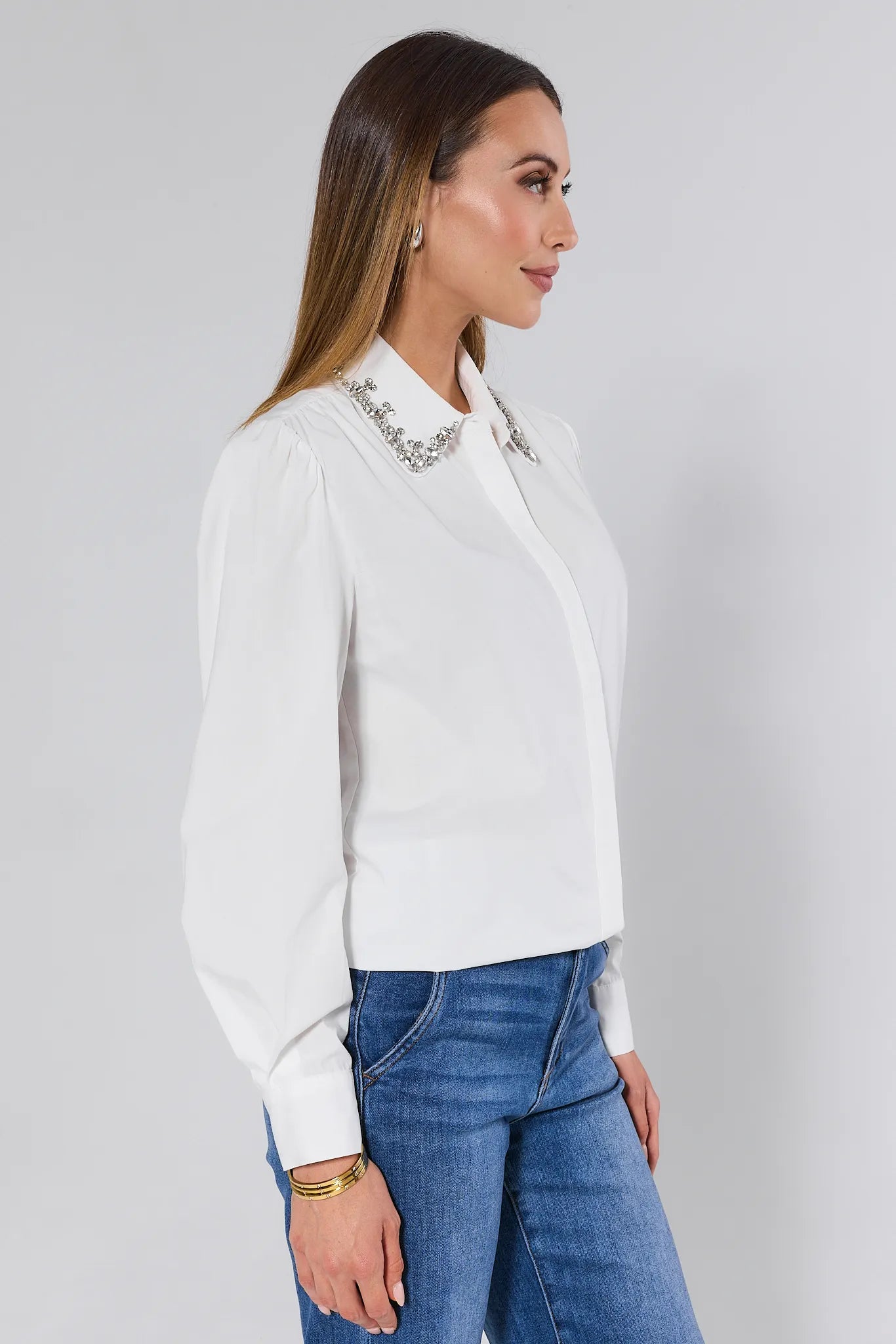 Woman wearing a white Celeste Top with embellishments and blue jeans on a plain background