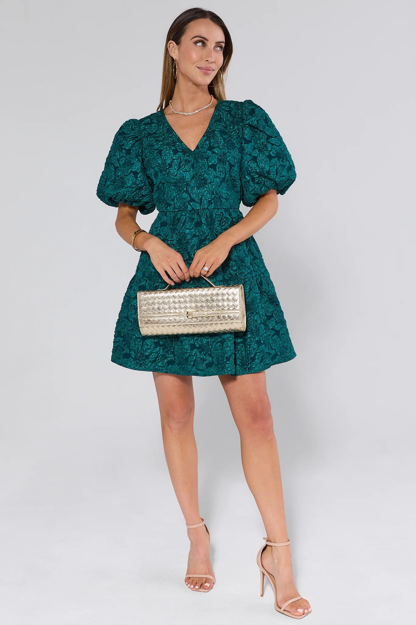 Woman wearing a green floral dress holding a silver clutch against a white background
