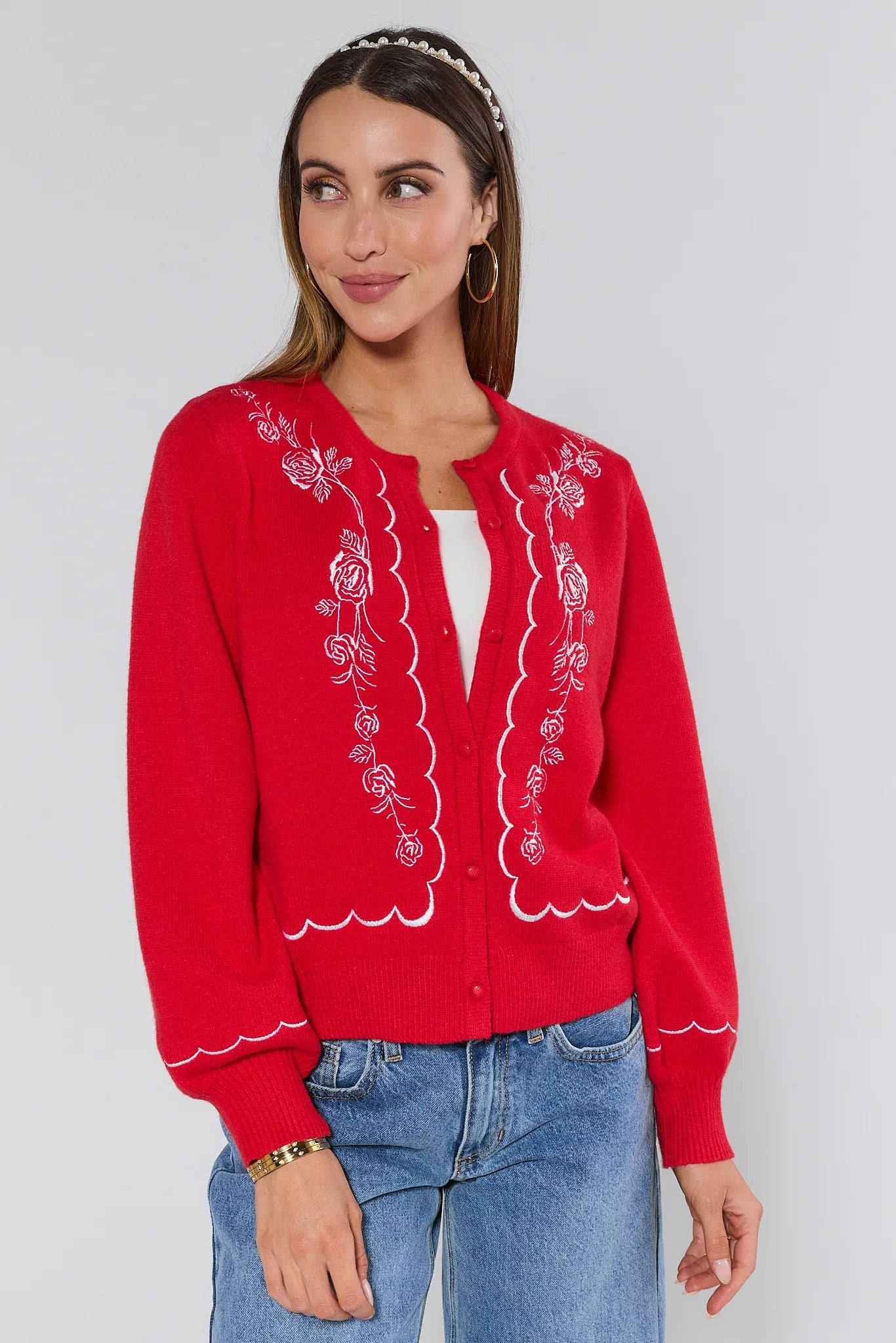 Woman wearing a red embroidered Ginger cardigan over a white top and blue jeans on a plain background