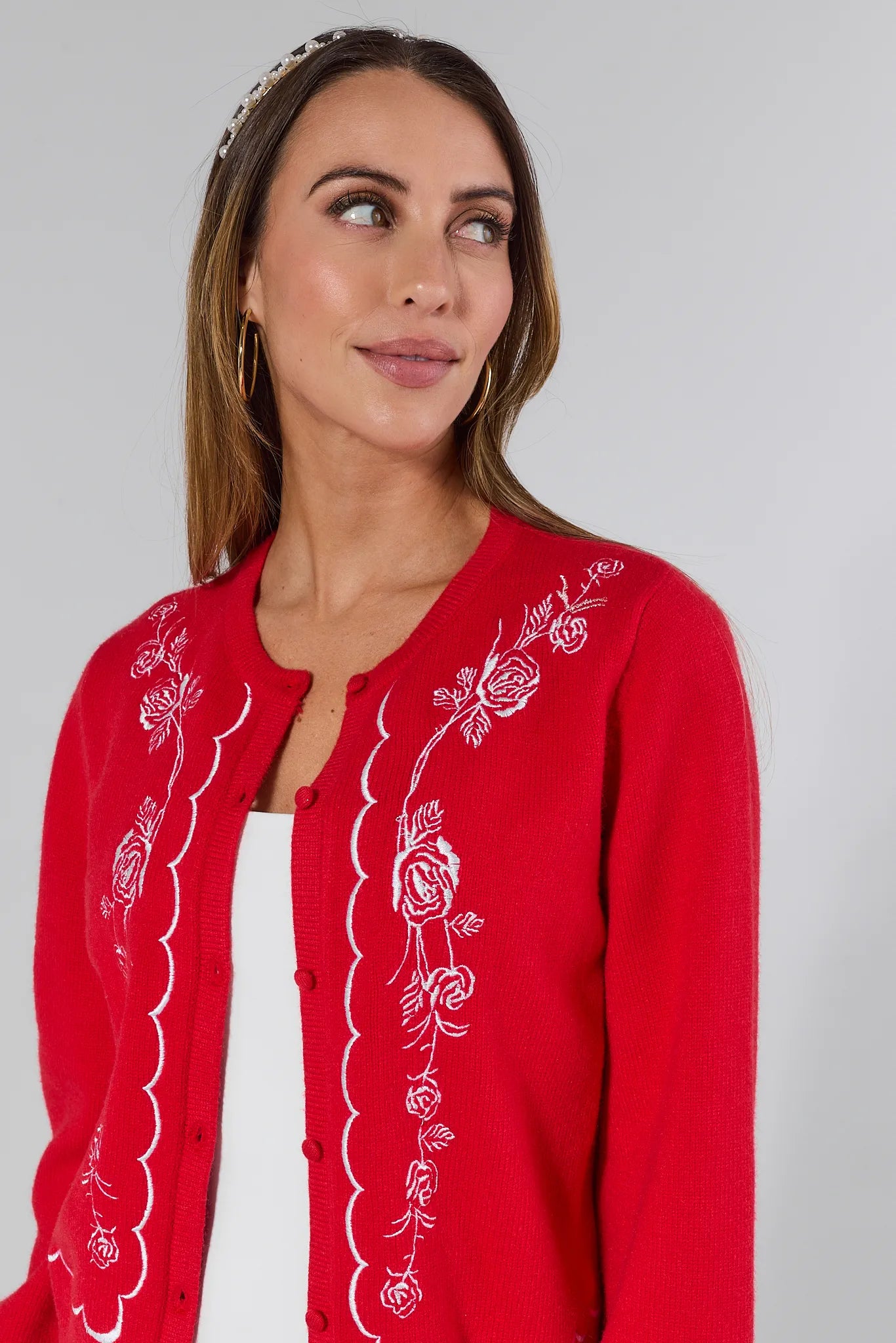 Woman wearing a red embroidered Ginger cardigan over a white top and blue jeans on a plain background
