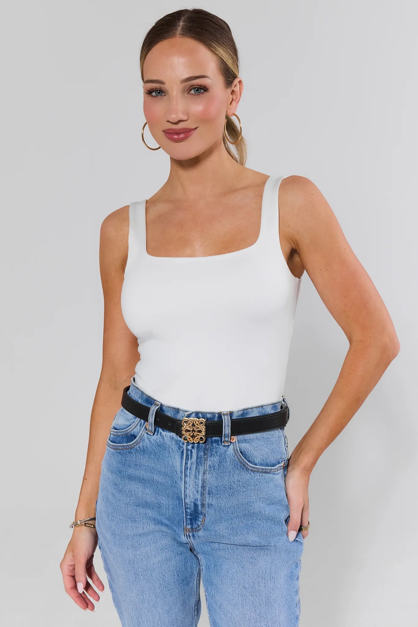 Woman wearing a white tank top and blue jeans with a black rowan belt against a plain background