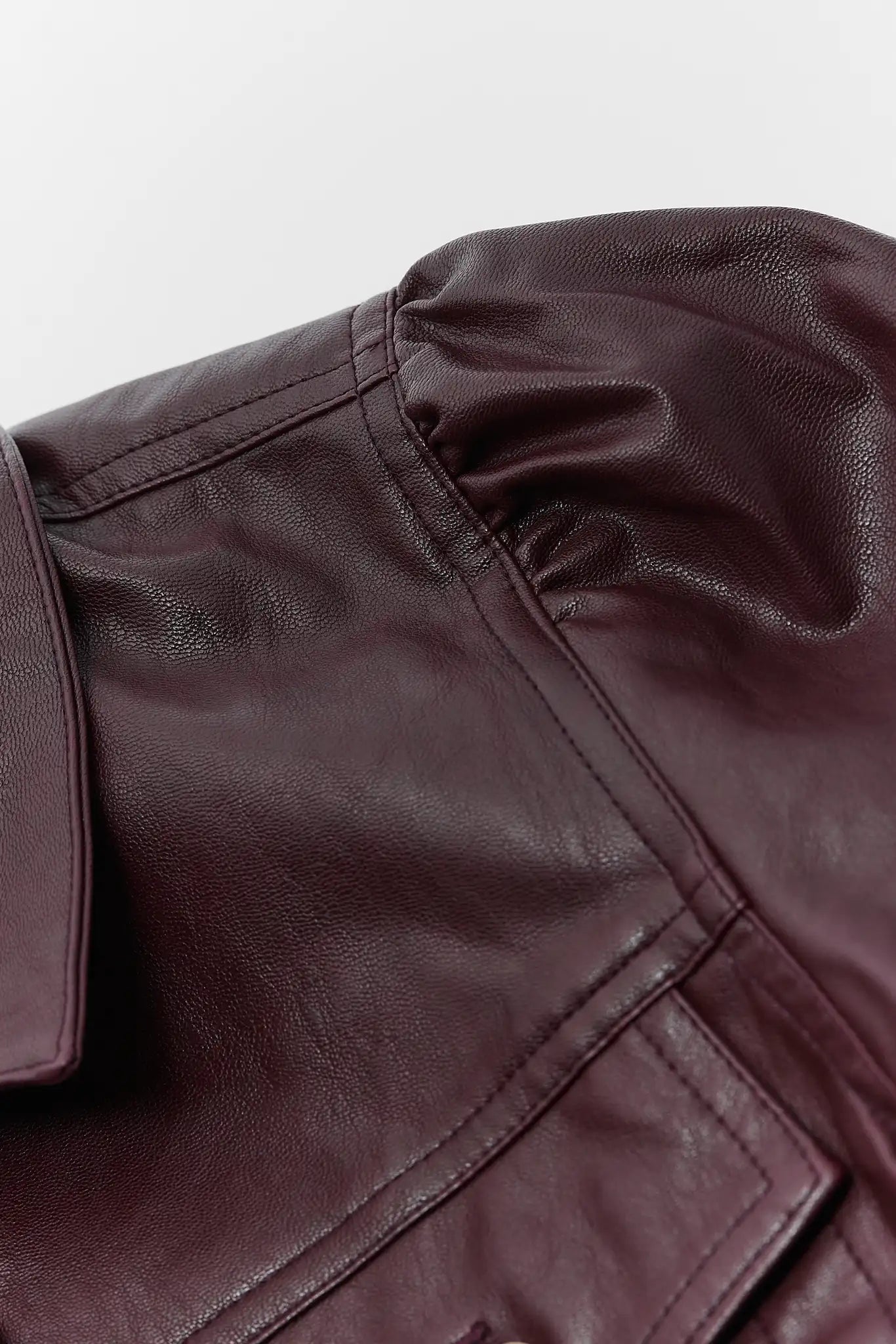 Close-up of a dark brown faux leather jacket showing the texture, stitching, and collar detail against a light gray background.