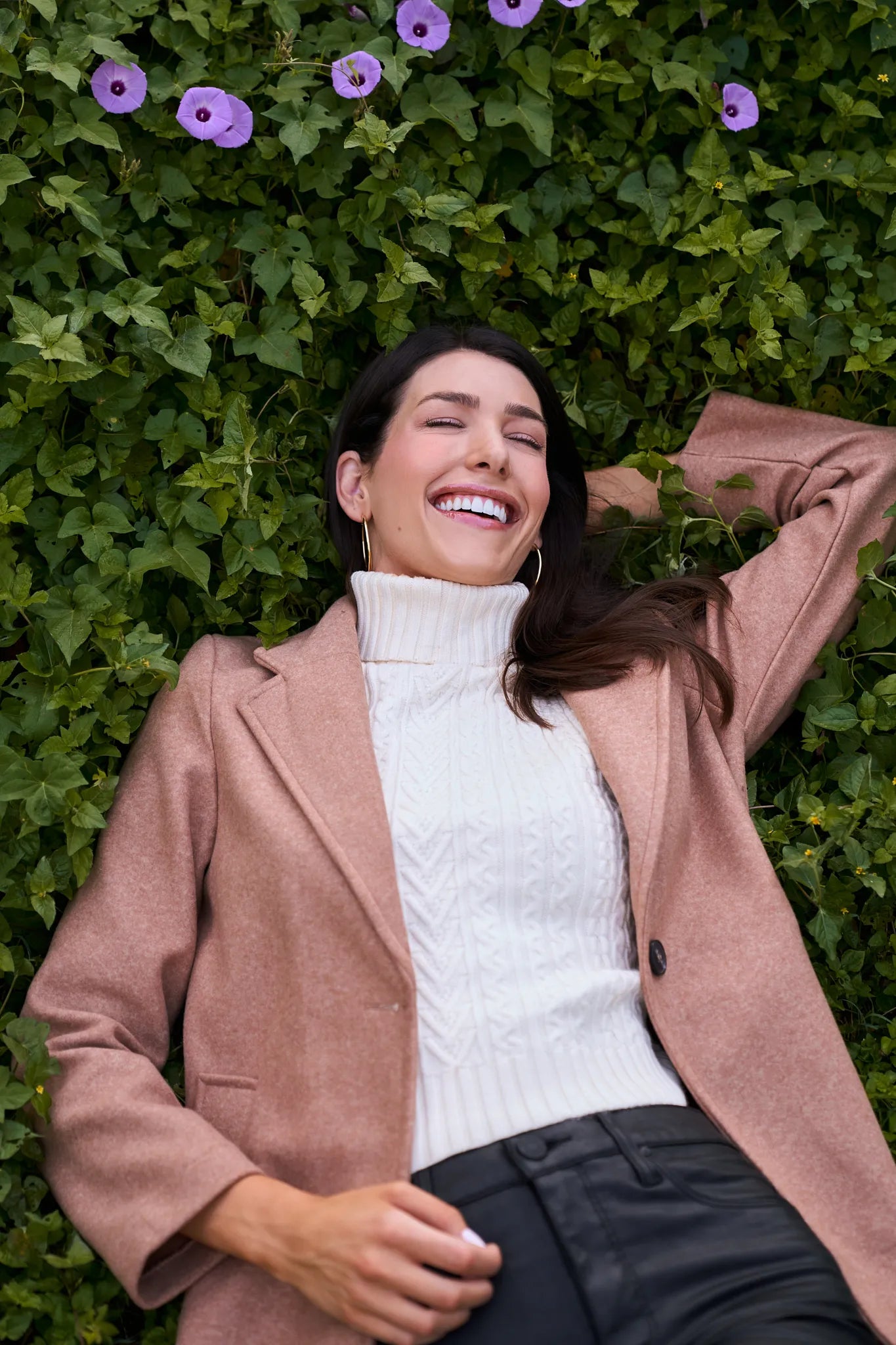 Woman in a Lauren Coat and white sweater standing against a green wall with purple flowers.