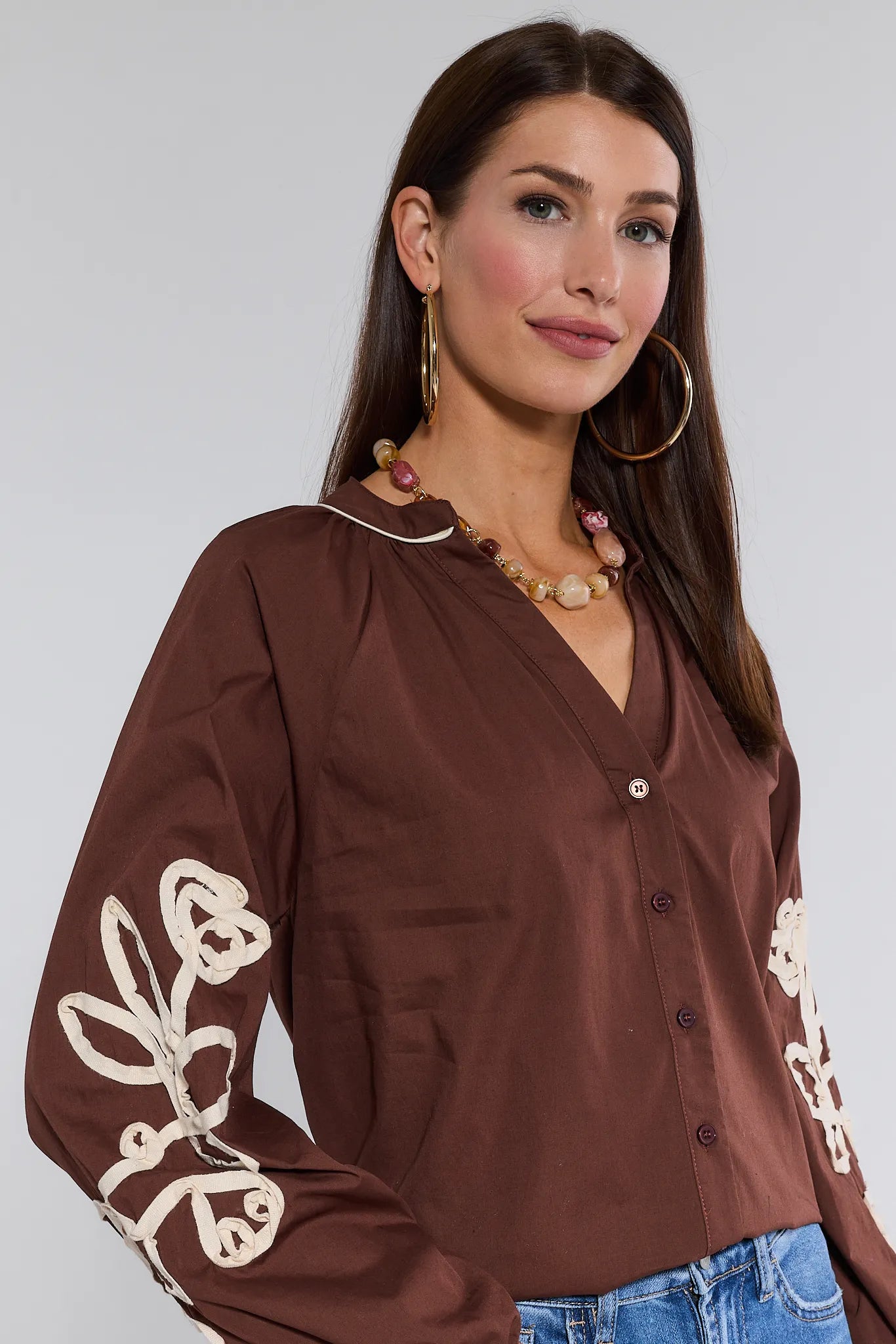 Woman wearing a brown Quinlan Top with white embroidery on sleeves against a plain background