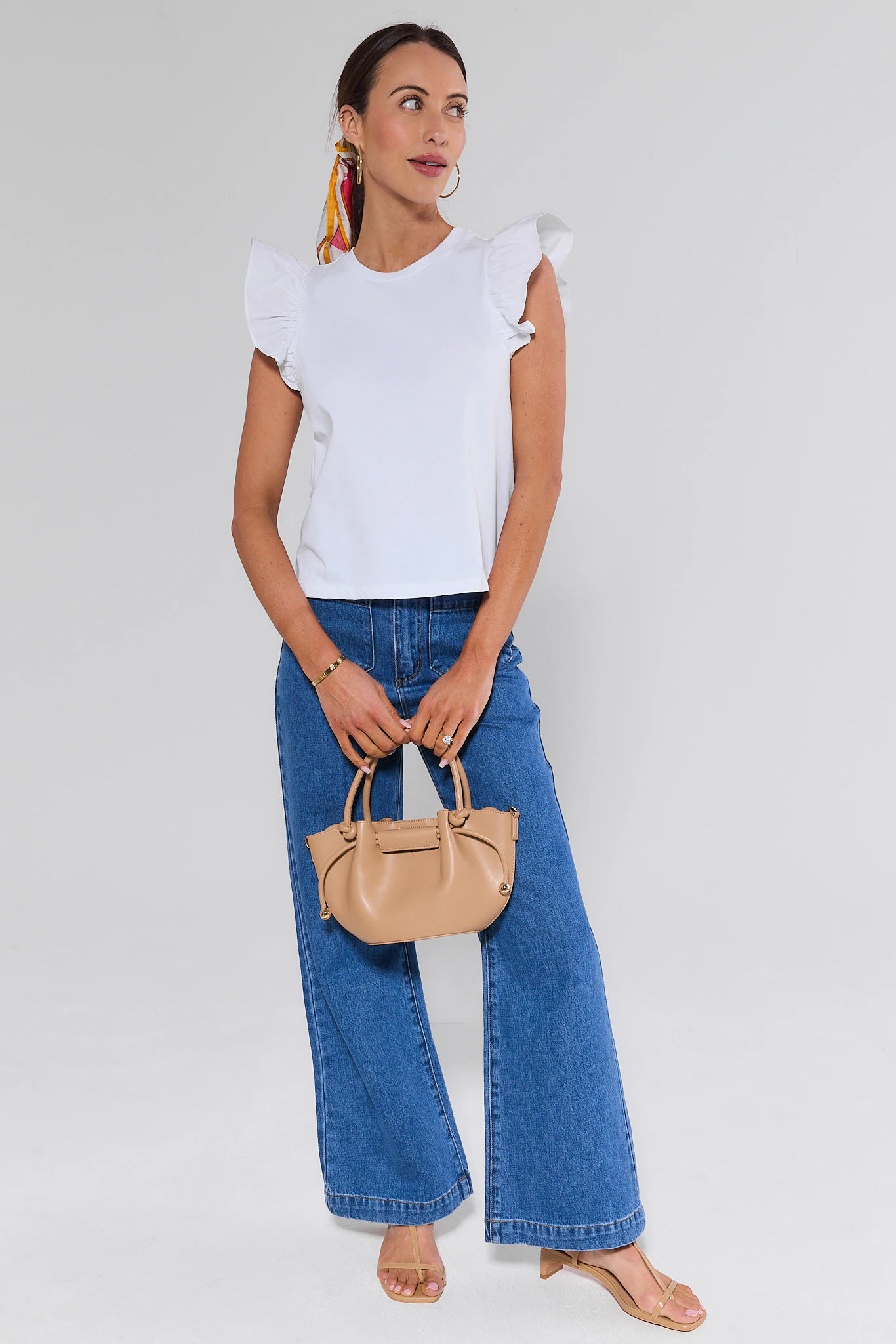 Woman wearing a white top and blue jeans, holding a beige handbag on a plain background