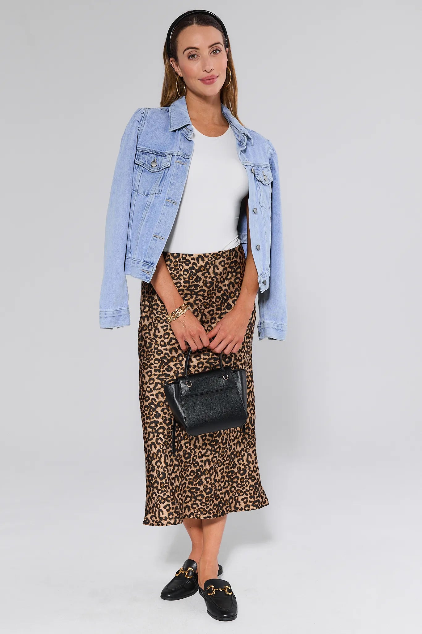 Woman wearing a denim jacket, white top, leopard print Gretchen Skirt, and black loafers on a plain background