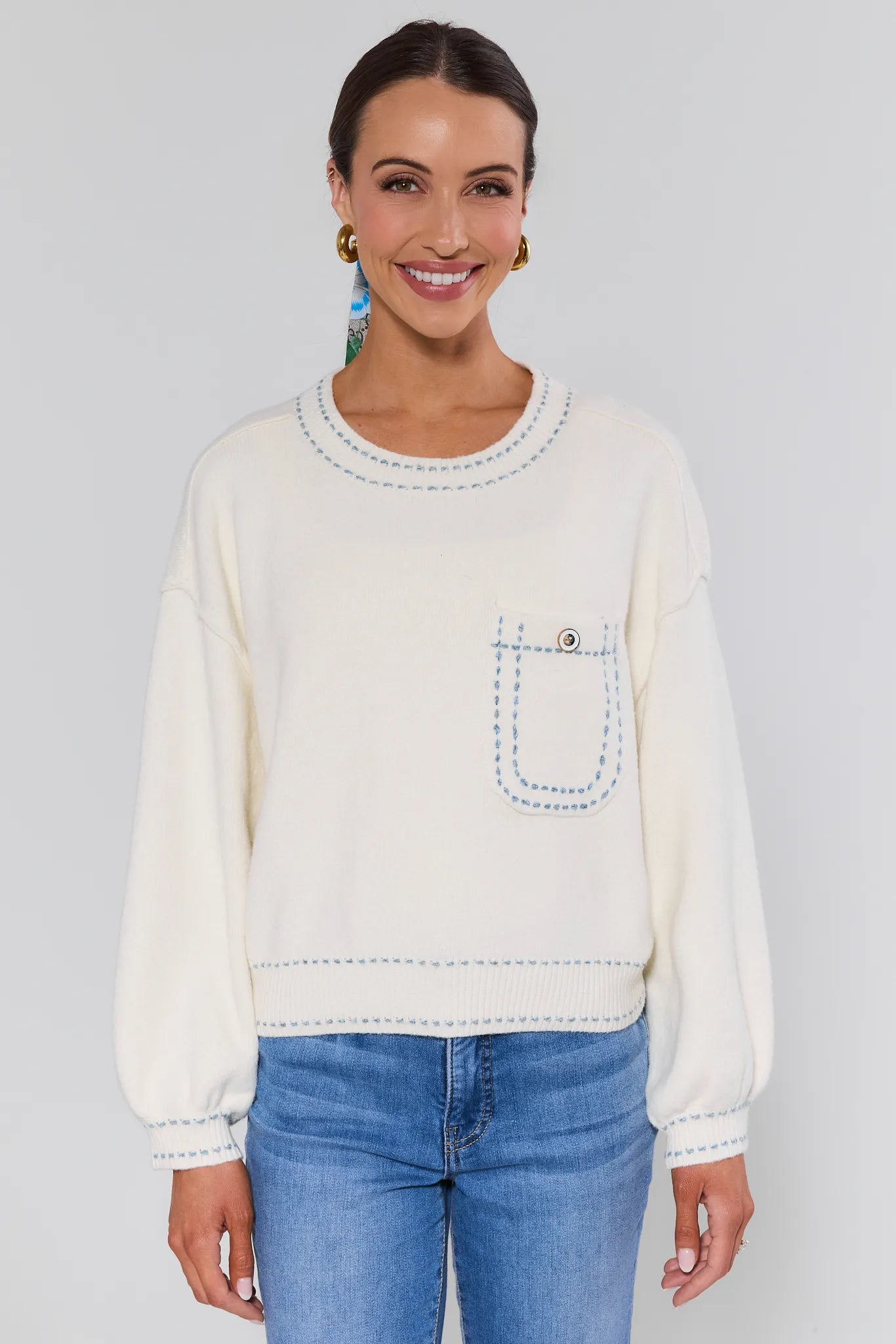 Woman wearing a cream sweater with a pocket and blue jeans on a plain background