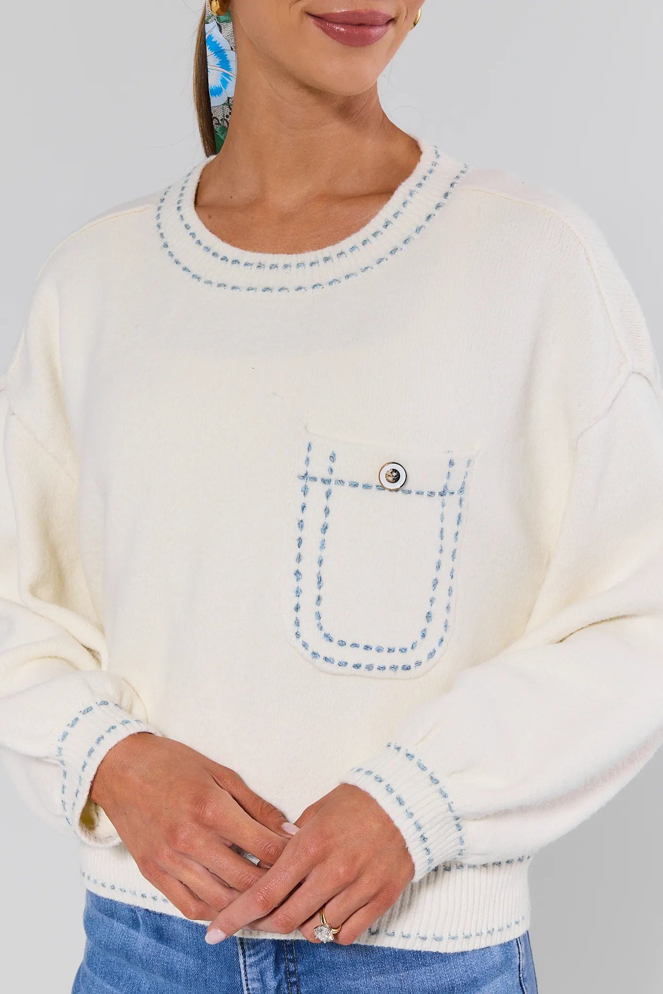 White sweater with blue stitching details worn by a person on a gray background