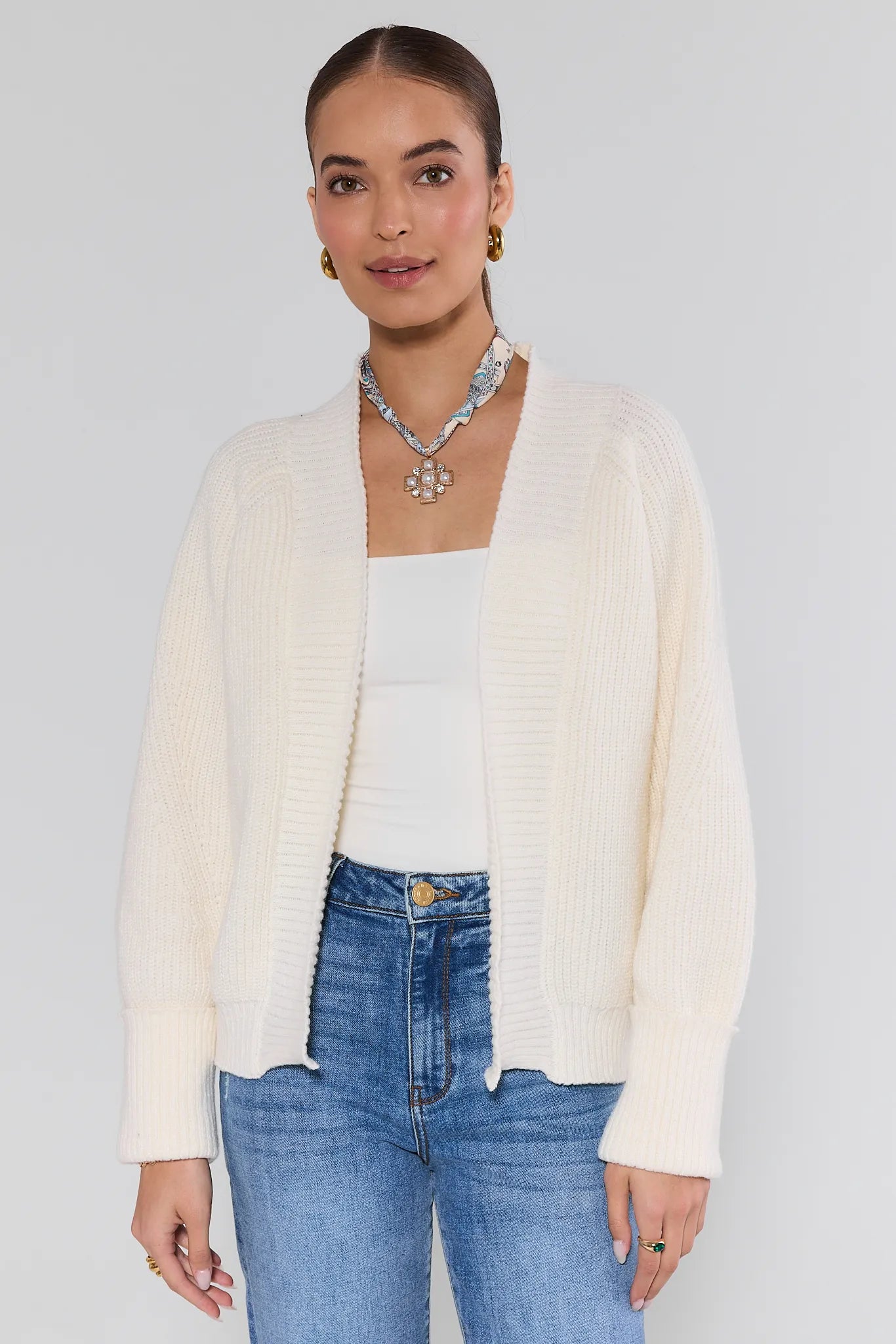 Woman wearing a cream cardigan, white top, and blue jeans against a plain background