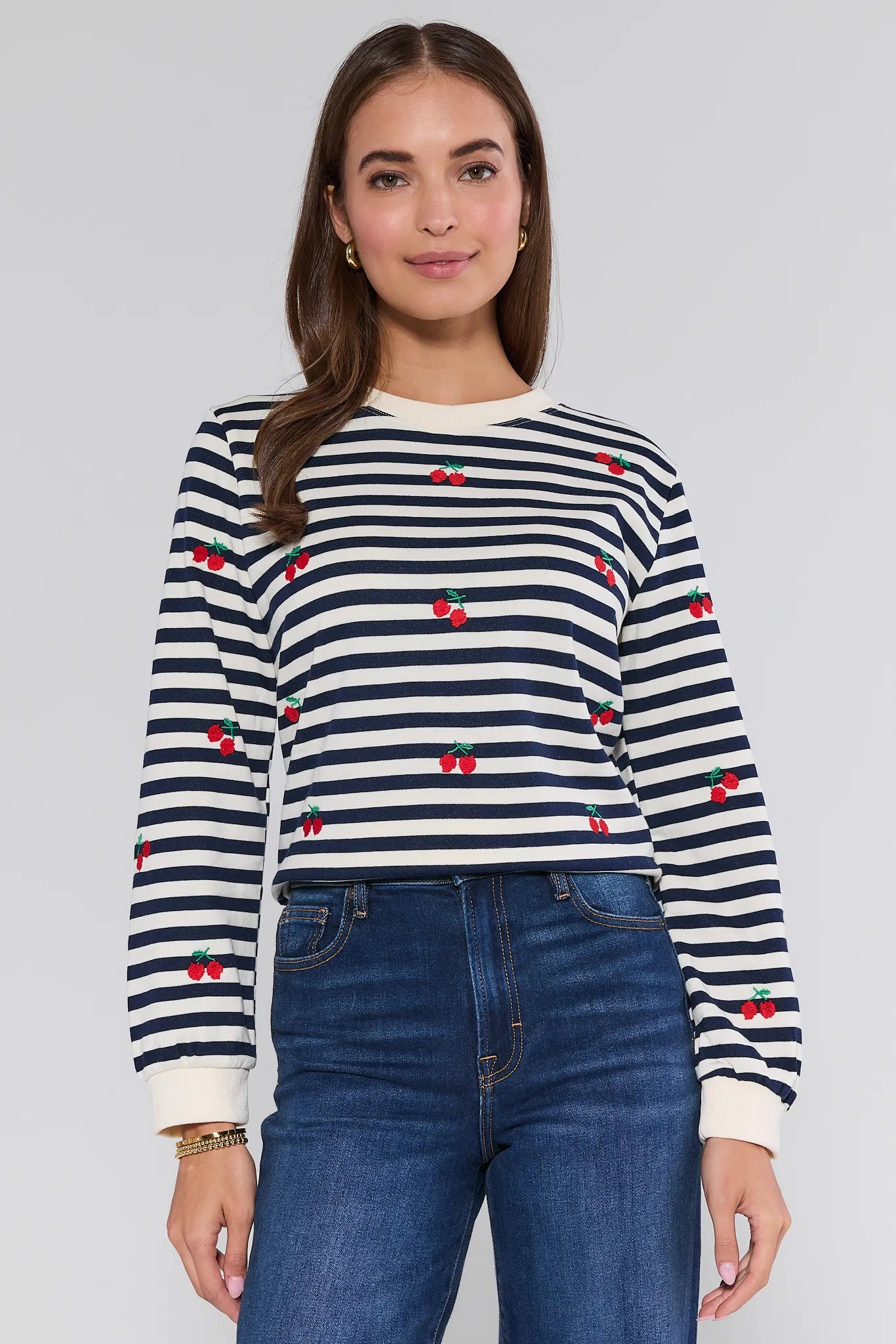 Woman wearing a striped Belle Sweatshirt with cherry patterns and blue jeans on a plain background