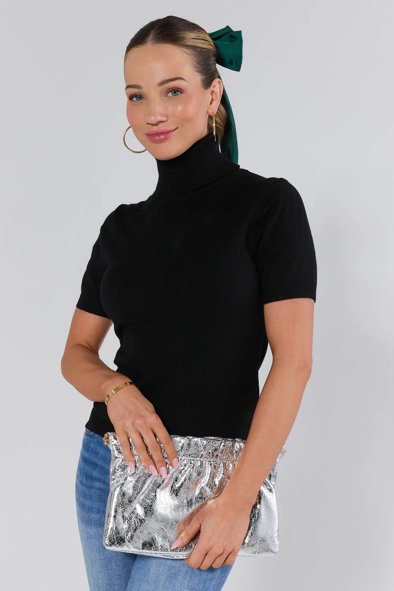 Woman wearing a black turtleneck and blue jeans, holding a silver clutch against a white background