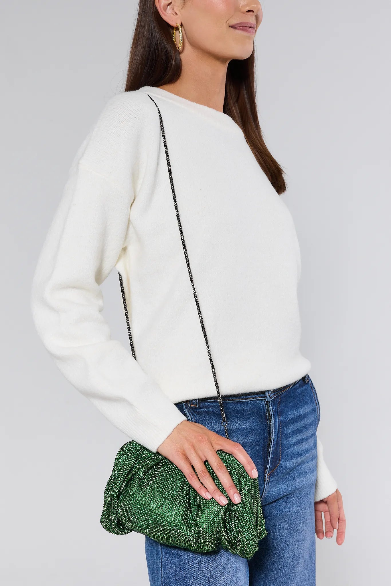 Woman wearing a white sweater and blue jeans, holding a green clutch against a plain background