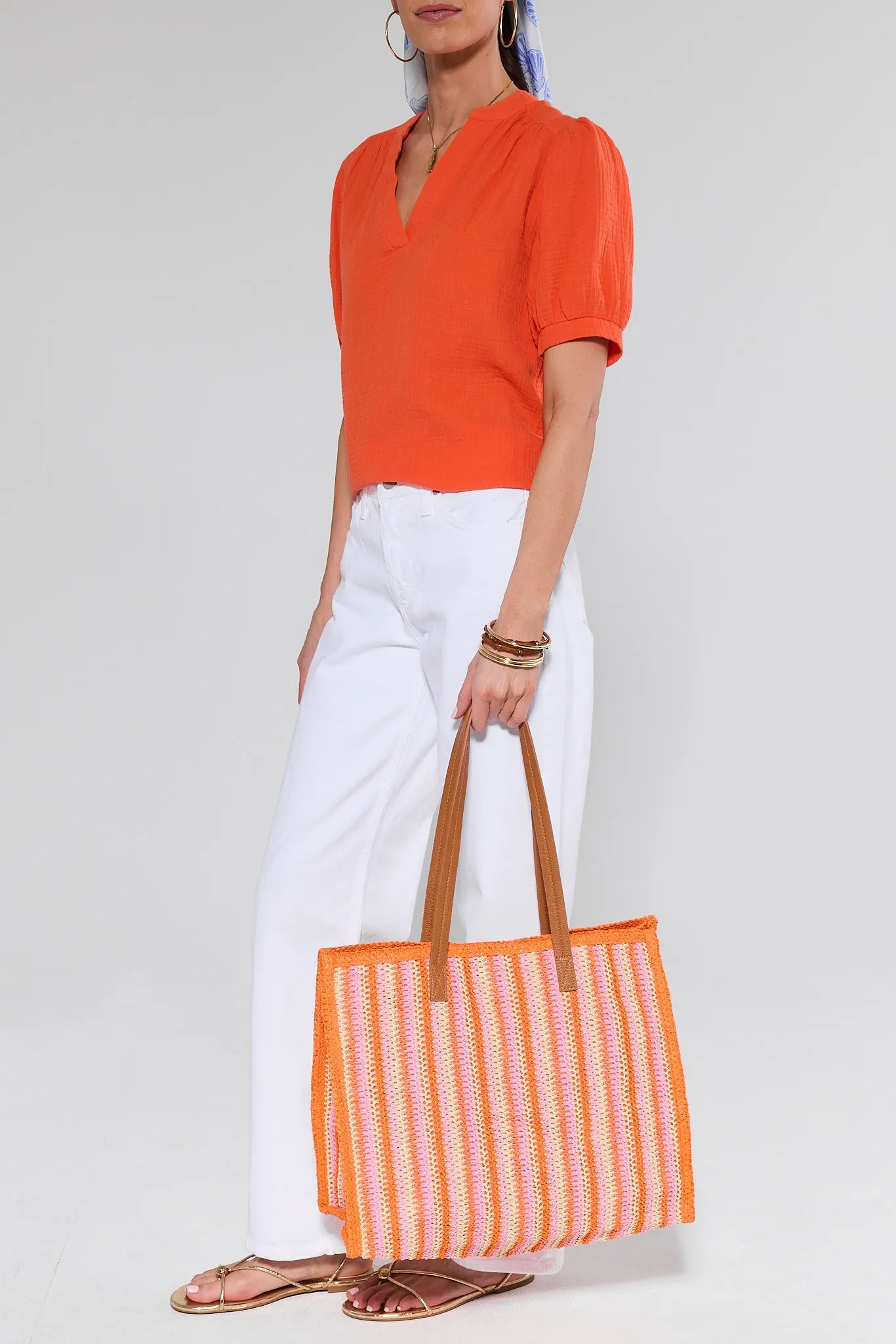 Person wearing an orange top and white pants, holding an orange and white striped bag on a light gray background
