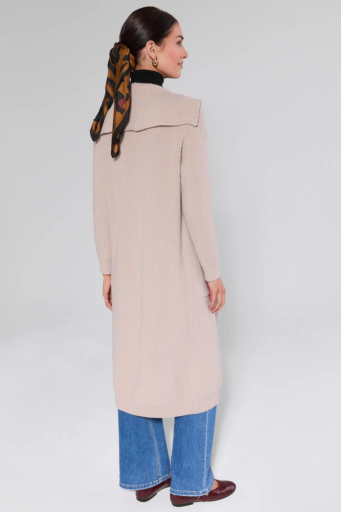 Woman wearing a Monroe Tan Knit Long Cardigan, black turtleneck, and blue jeans on a white background