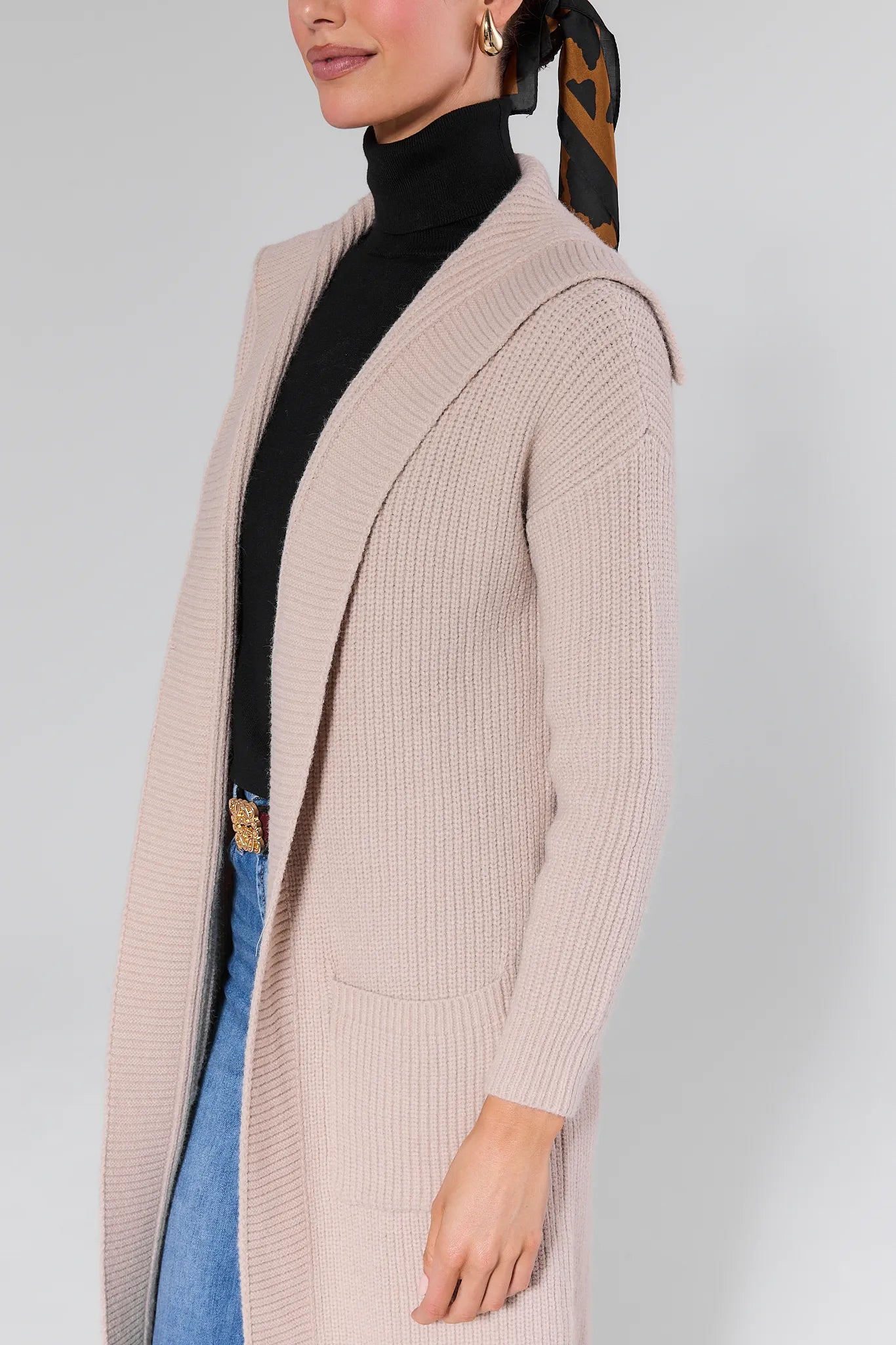 Woman wearing a Monroe Tan Knit Long Cardigan, black turtleneck, and blue jeans on a white background