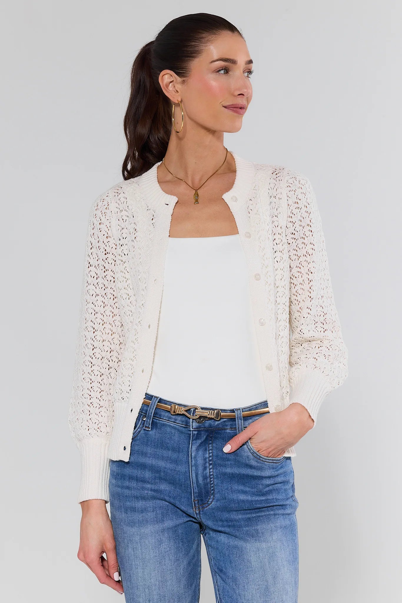 Woman wearing a white cardigan, white top, and blue jeans against a plain background