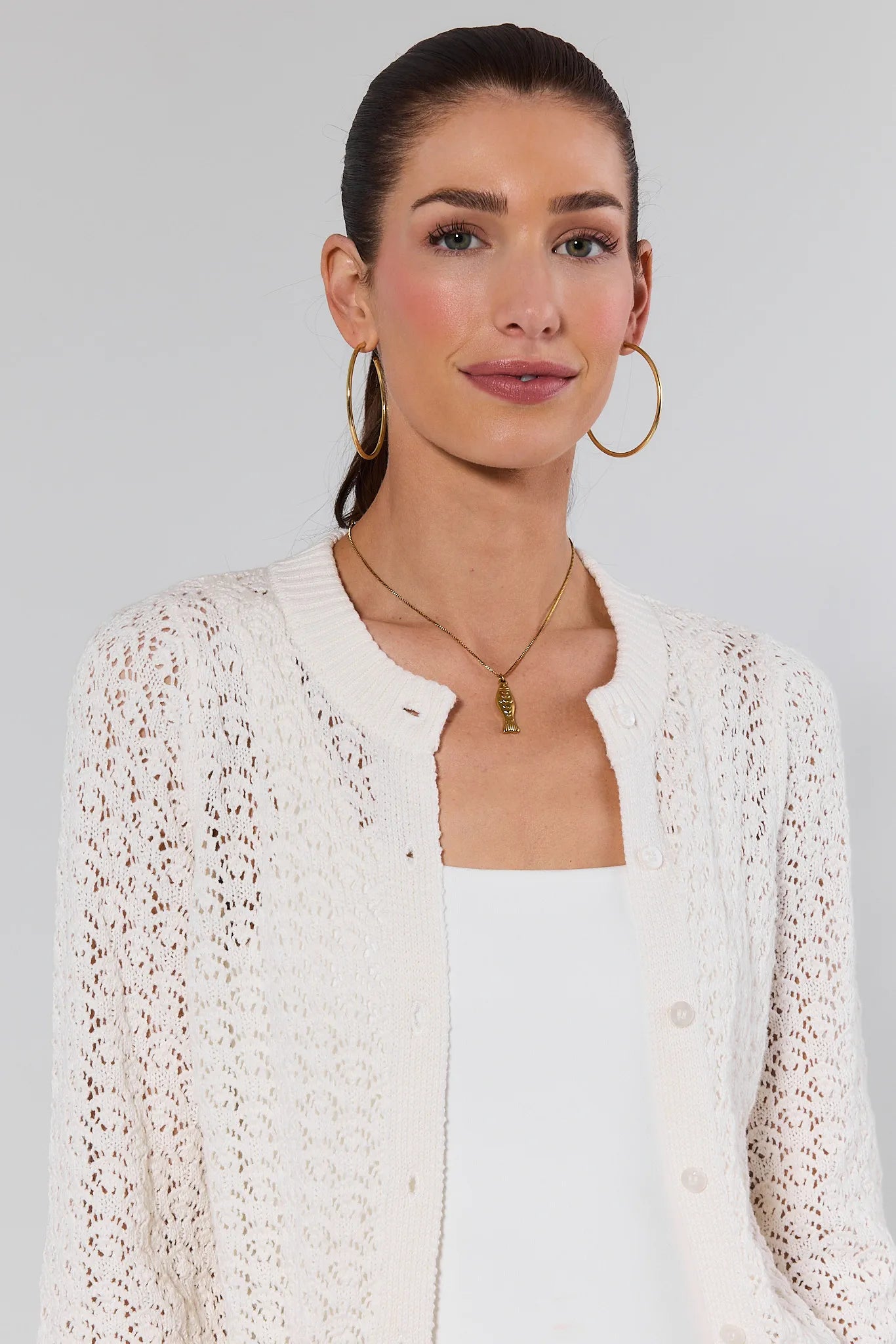 Woman wearing a white lace cardigan over a white shirt with gold hoop earrings and necklaces on a light gray background